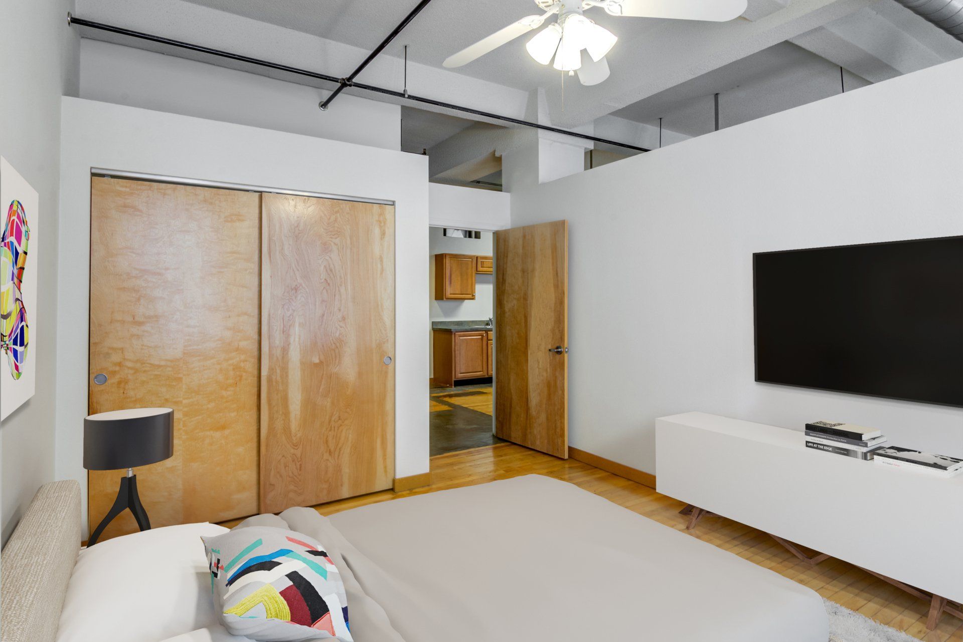 A modern, minimalist bedroom with a bed, wood closet doors, wall-mounted television, and a white console table.