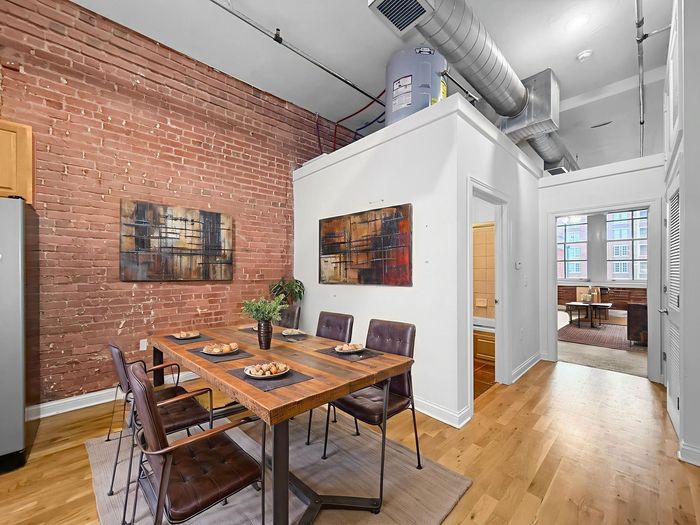 A modern dining room with a wooden table, brown leather chairs, and an exposed brick wall, leading into a living area.