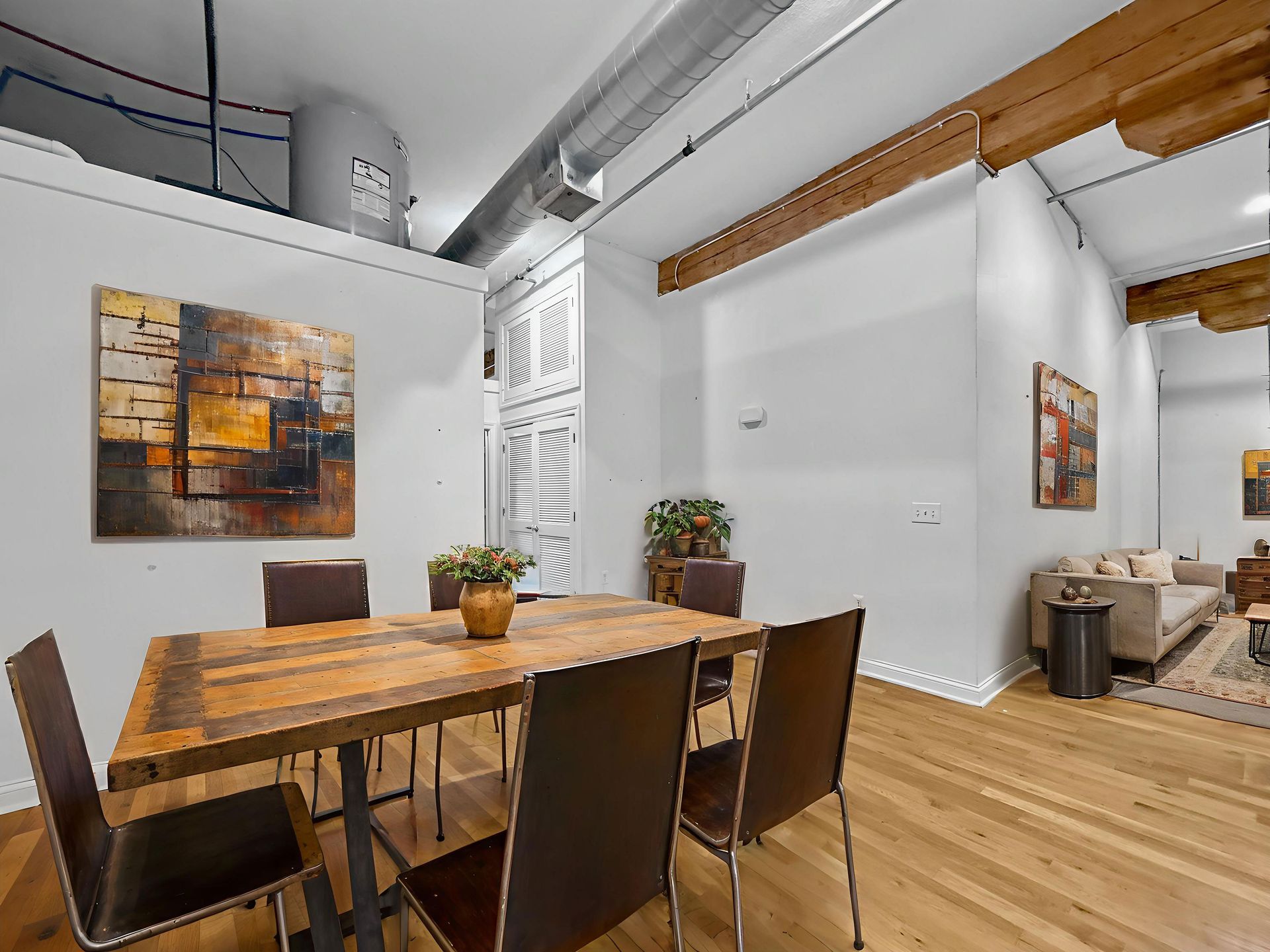 A dining area with a rustic wooden table and chairs on hardwood floors, opening to a living room with exposed beams.