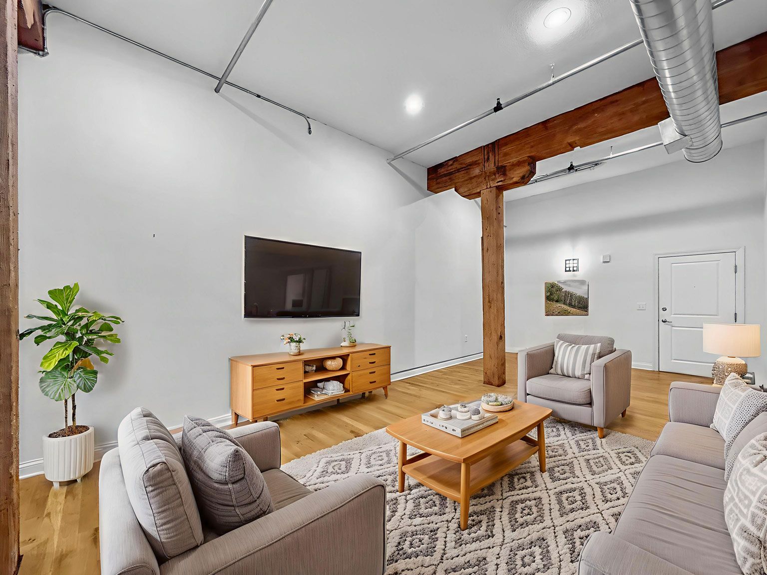Modern living room with a TV, light wood console, armchairs, sofa, patterned rug, and a rustic wooden support beam.