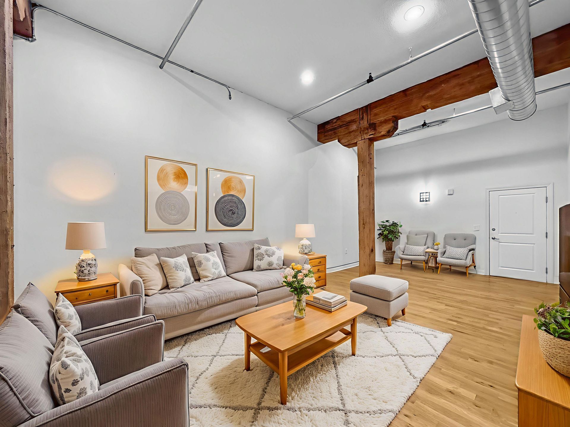 A bright, modern industrial-style living room with exposed wood beams, grey sofa, wood coffee table, and white rug.