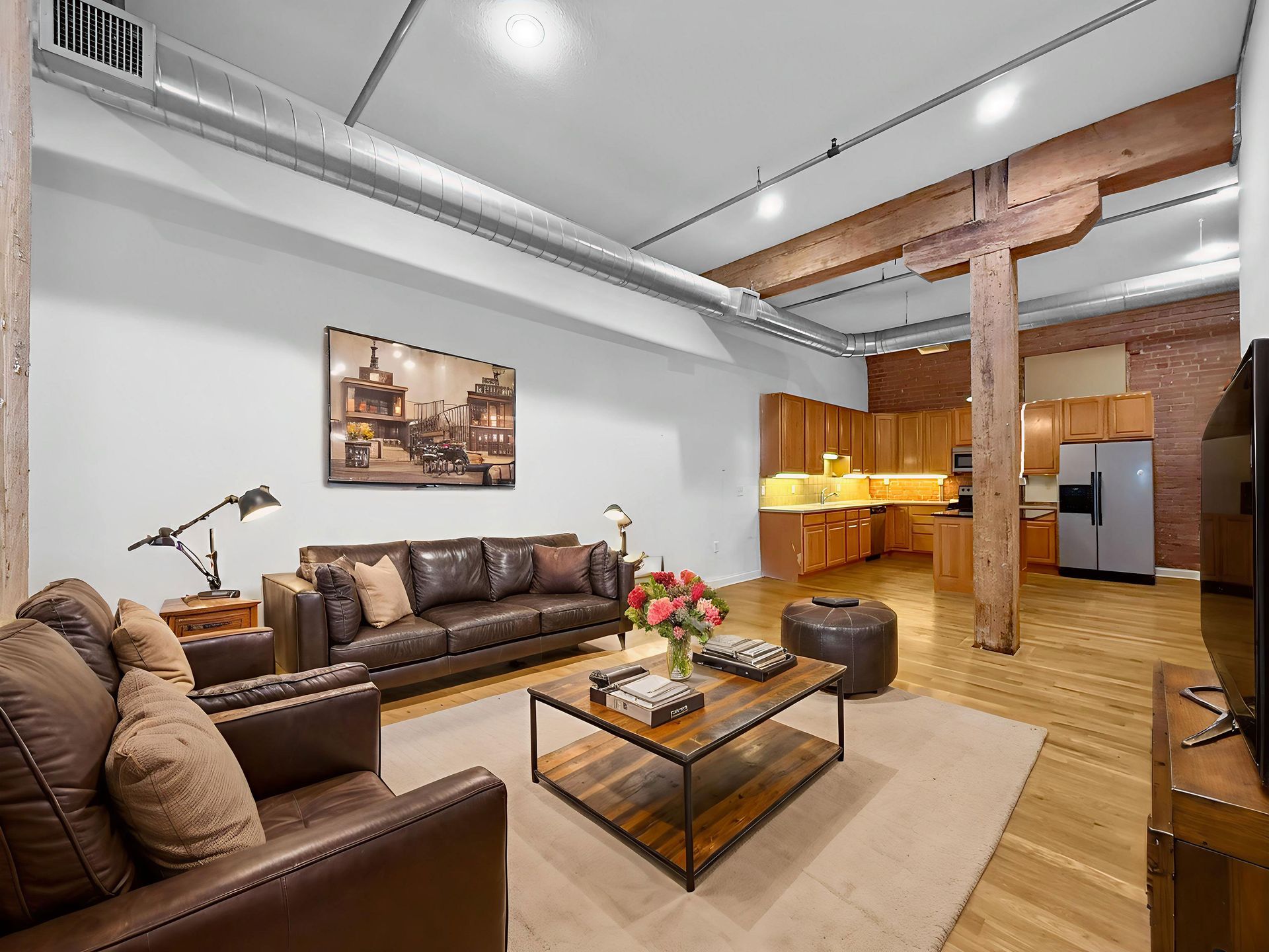 An open-plan loft living room with a brown leather sofa set, coffee table, and exposed wooden beams above a kitchen area.