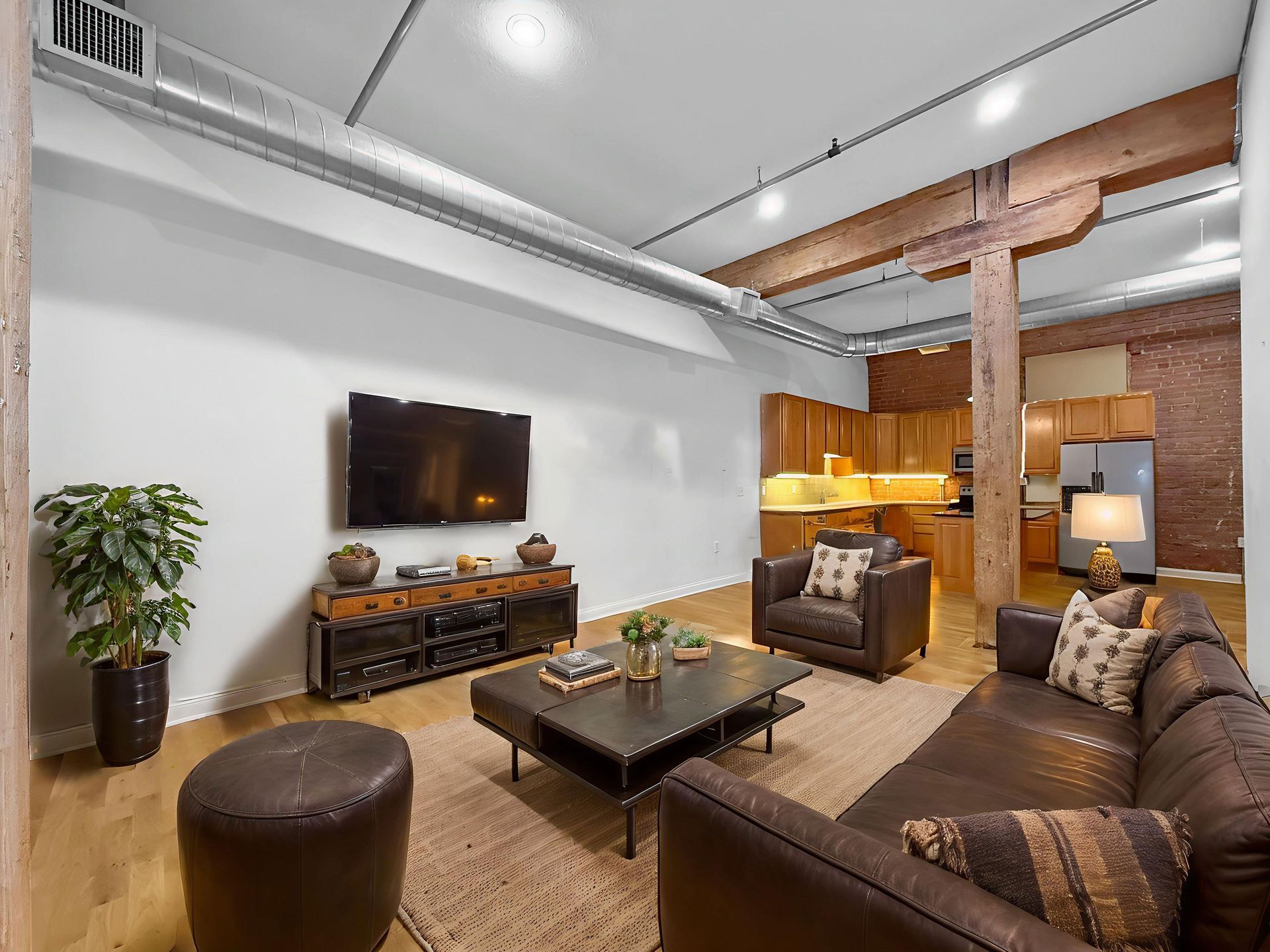 Living room with a leather sofa, armchair, coffee table, and TV, featuring exposed brick walls and an open-concept kitchen.