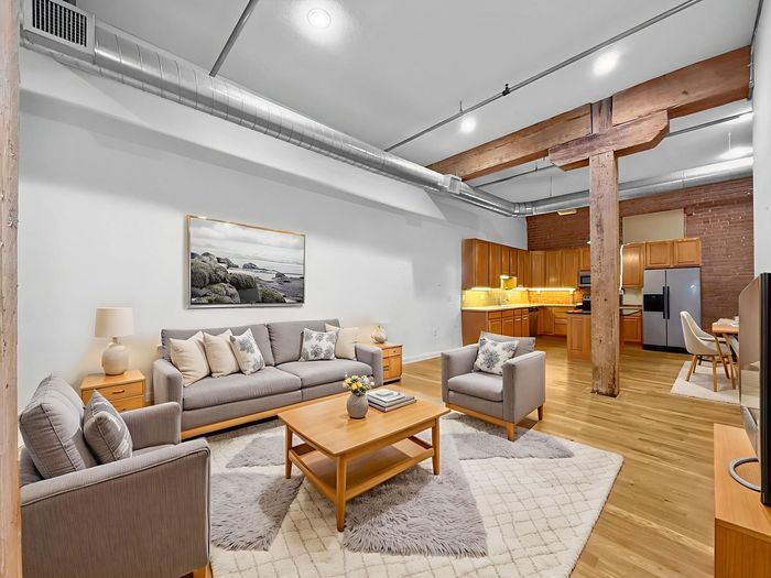 A modern loft living room with a grey sofa, armchair, coffee table, and an open-concept kitchen with wood cabinets.
