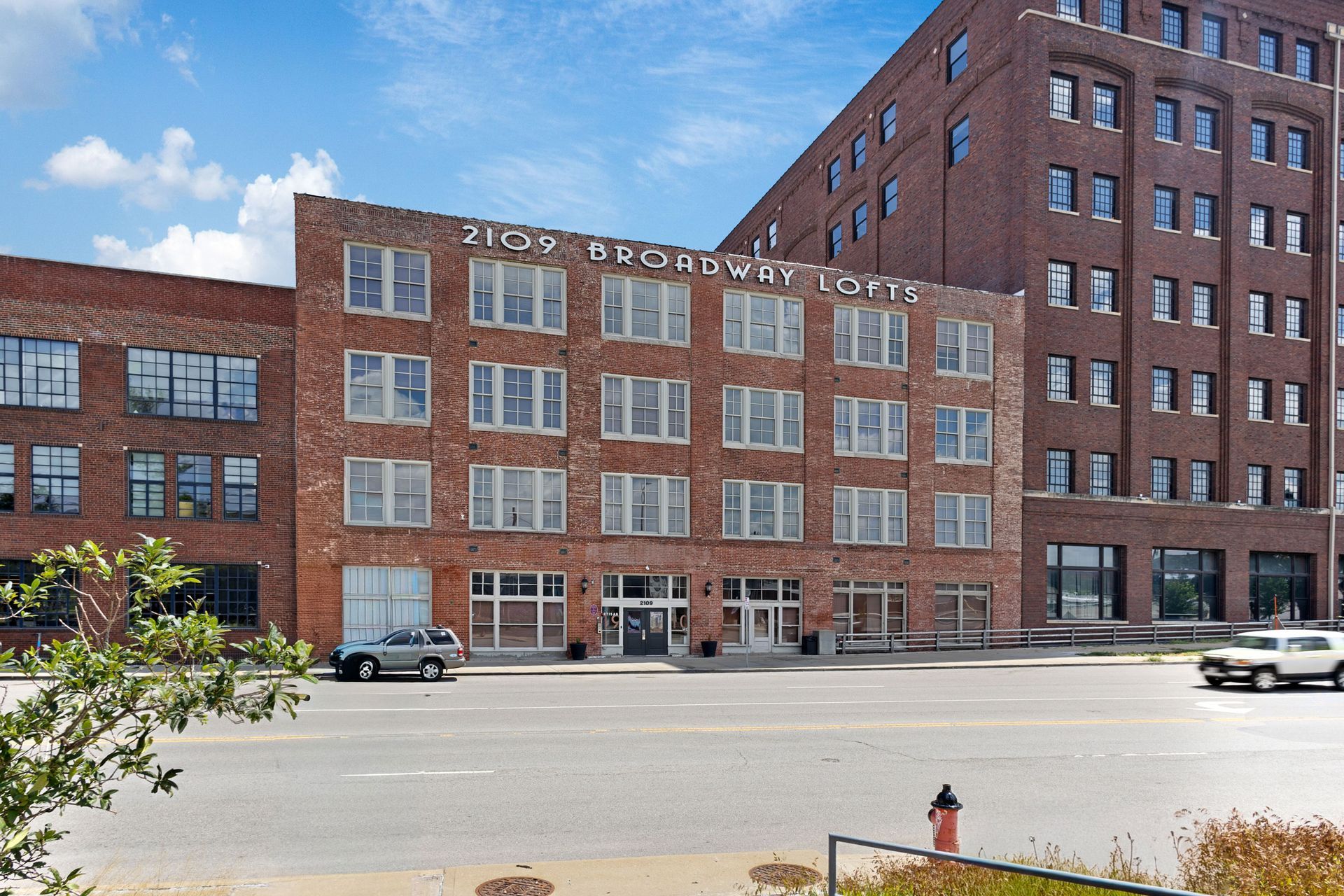 Red brick commercial buildings with large windows line a wide city street under a blue sky with scattered clouds.