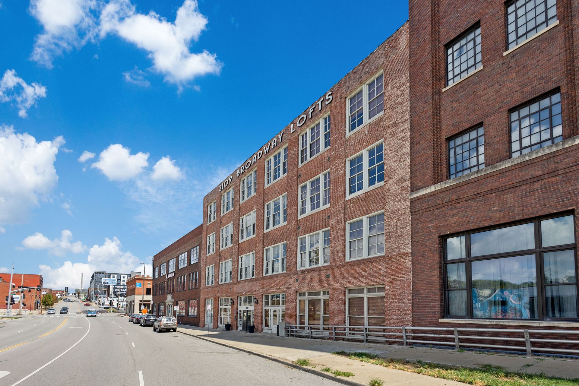 A street-level view of a multi-story red brick commercial building under a bright blue sky with scattered clouds.