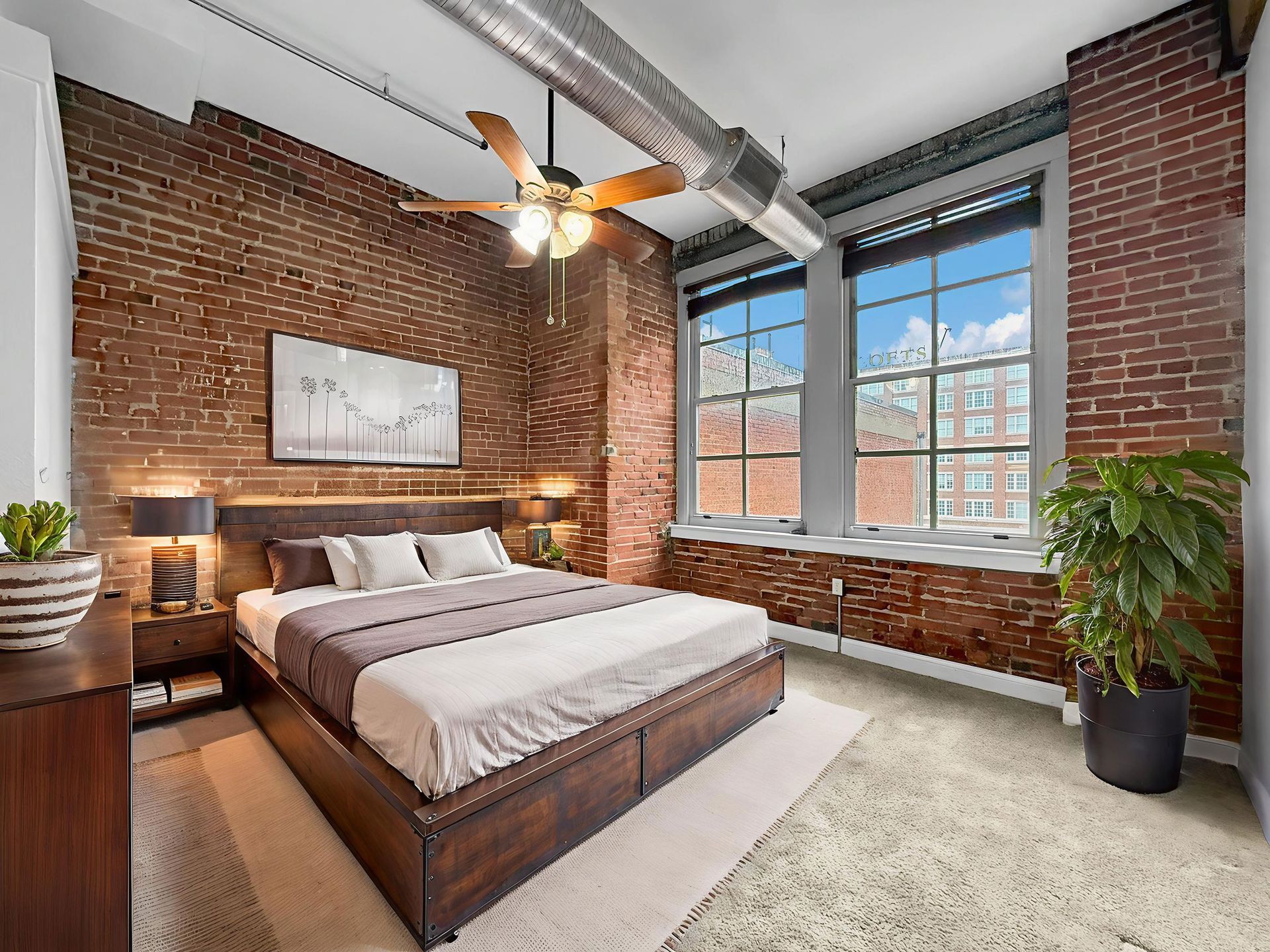 Modern industrial loft bedroom with brick walls, a wooden bed, large windows, and a green plant.
