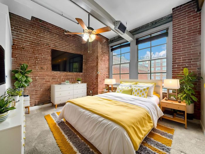 A bedroom with exposed brick walls, a queen bed with yellow bedding, large windows, and a ceiling fan.
