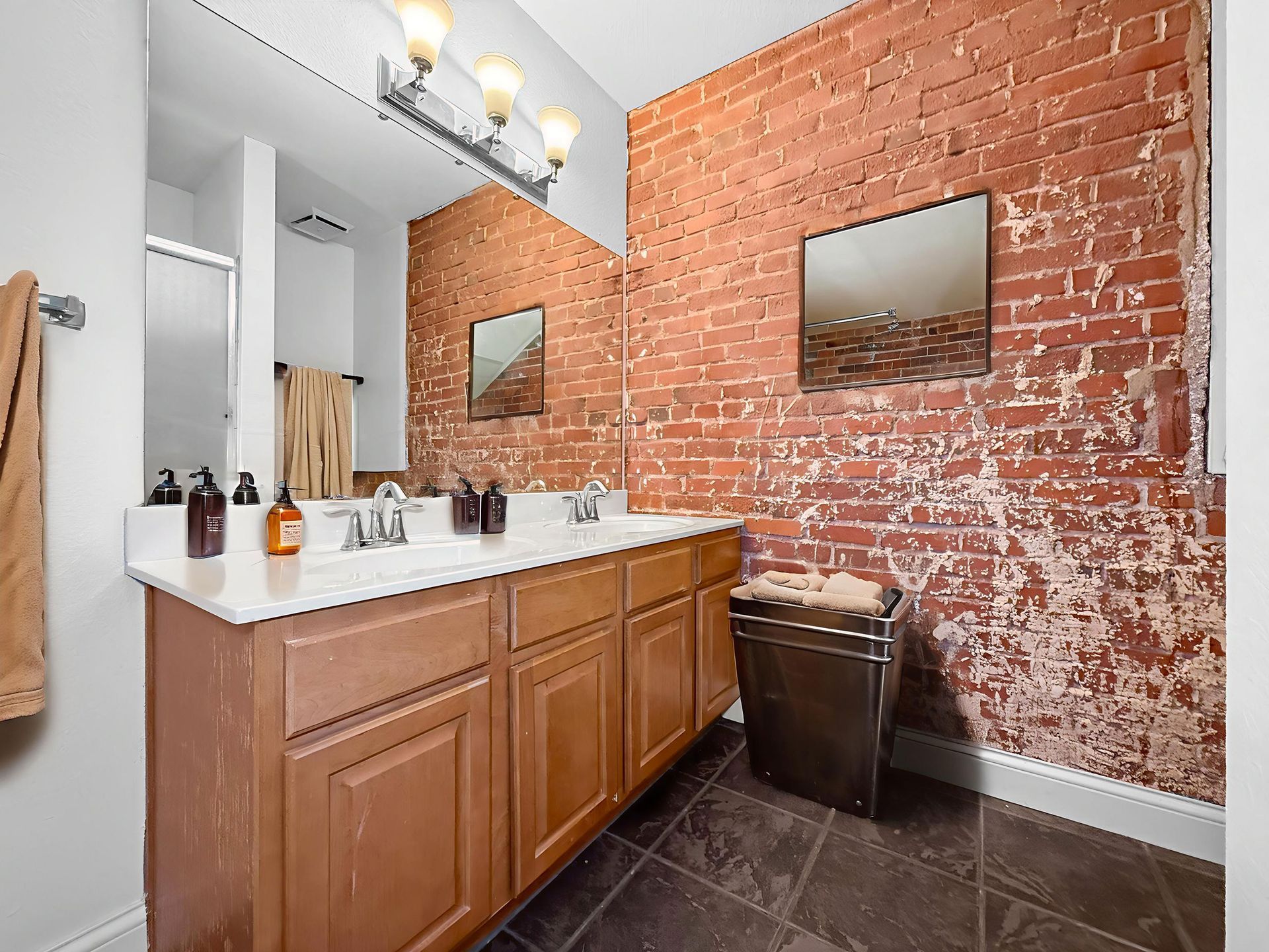 A bathroom vanity with a light countertop, wood cabinets, and a mirror against an exposed red brick wall with tile floors.