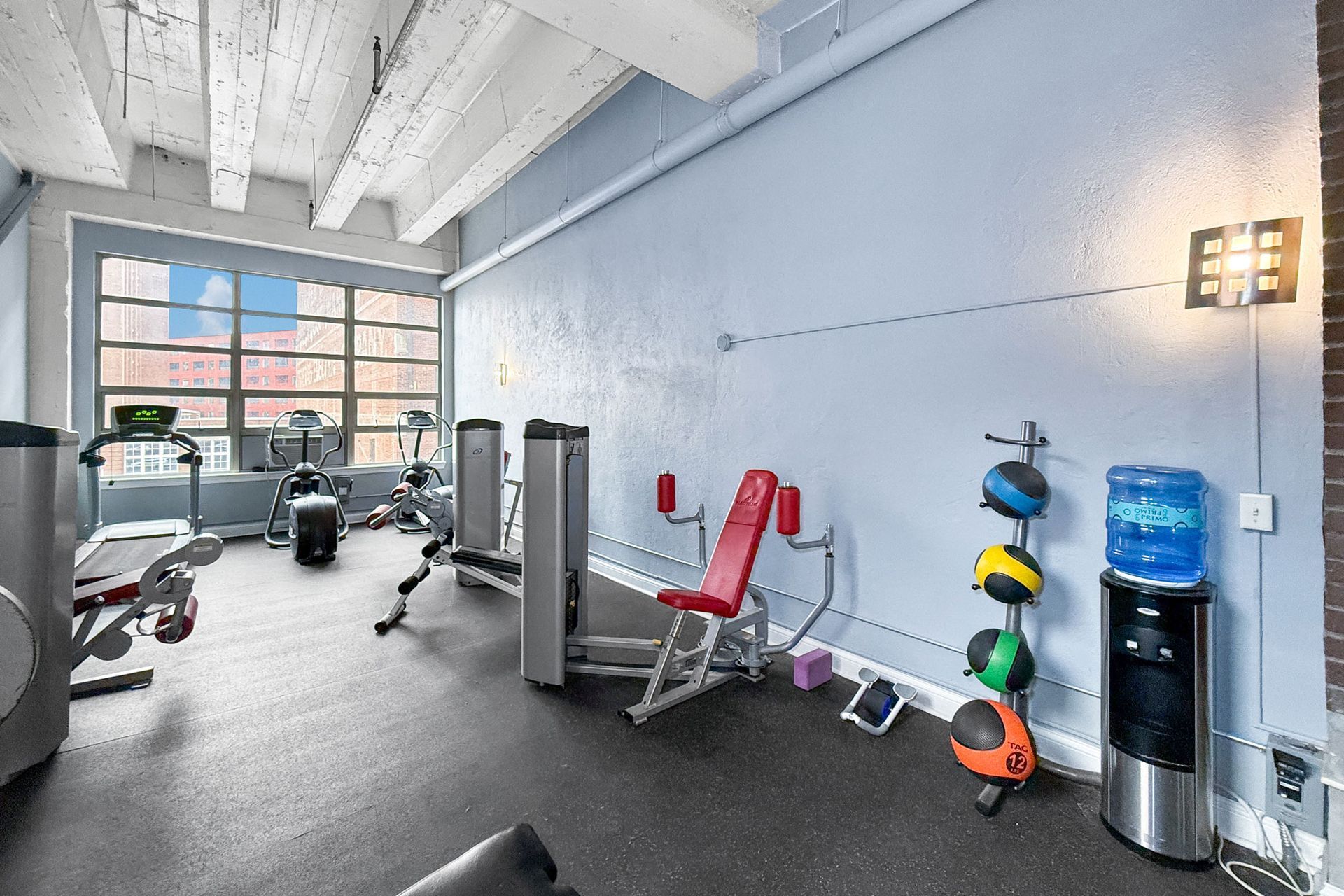 A small, brightly lit gym with exercise machines, a red weight bench, a medicine ball rack, and a water cooler.
