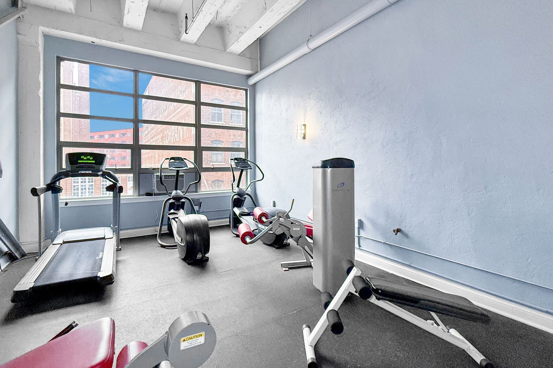 A small fitness center with a treadmill, elliptical, and strength training equipment against a light blue wall.