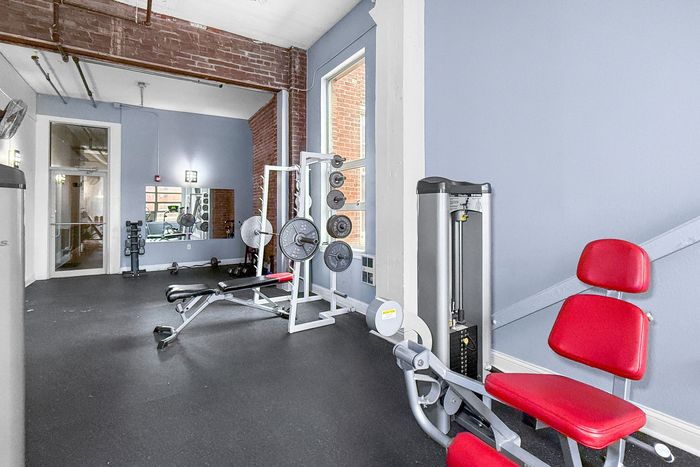 A small gym with a weight machine featuring a red seat, a weight bench, and a rack with weight plates on a black floor.