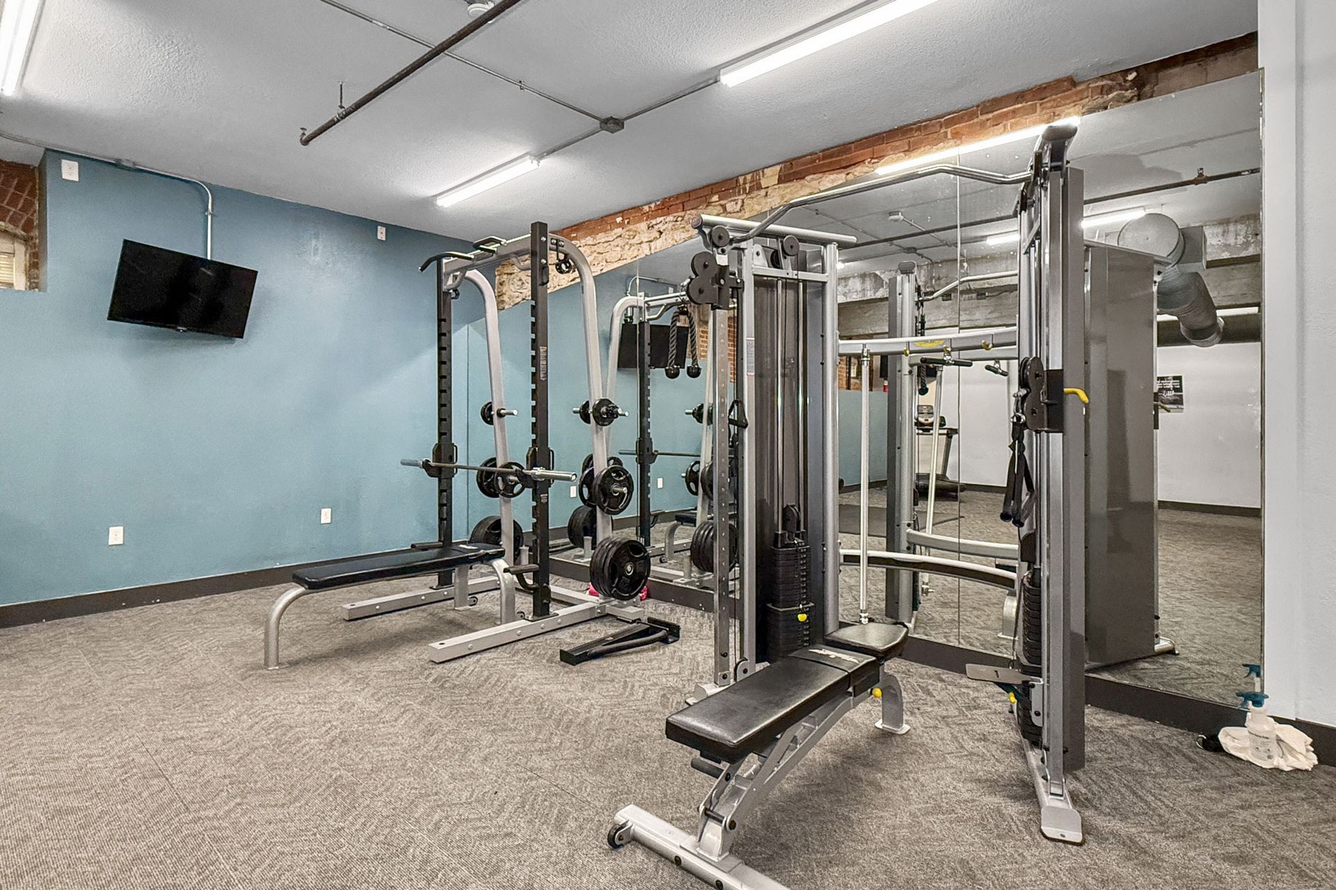 An indoor gym featuring a squat rack and a cable machine with benches on gray carpeted flooring.