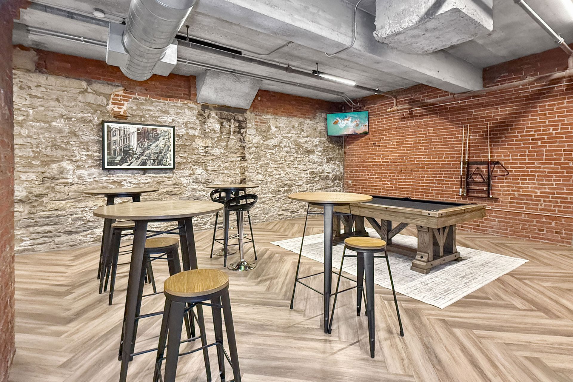 A rustic bar area featuring high wooden tables, stools, a pool table, stone and brick walls, and exposed ceiling pipes.
