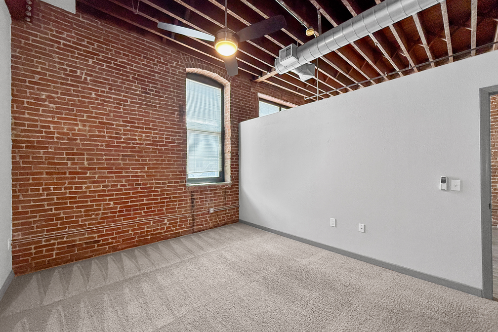 An unfurnished room featuring an exposed brick wall, a window, a ceiling fan, and grey carpet flooring.