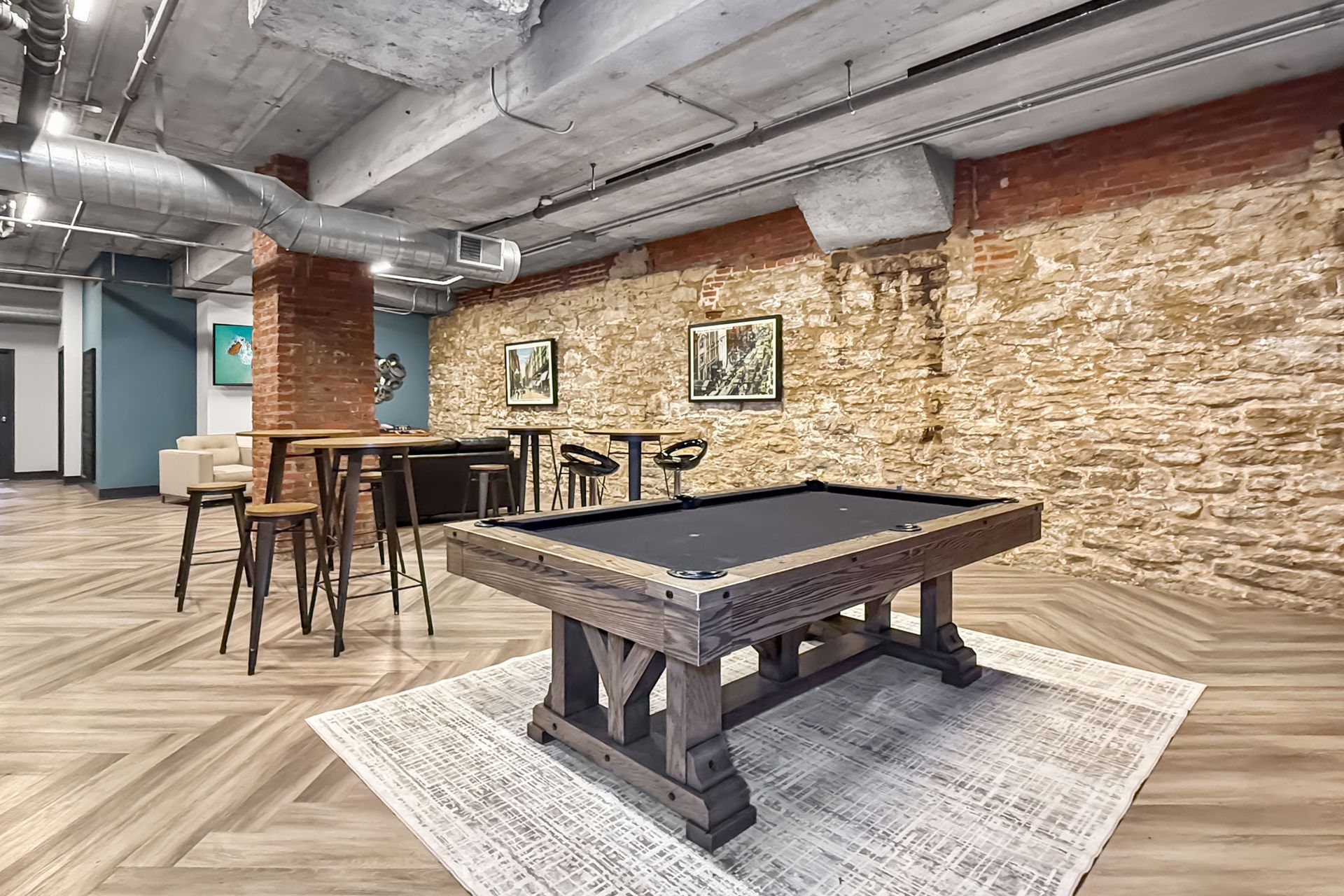 A pool table on a gray rug in a modern lounge with exposed brick walls, industrial ceiling, and wood-patterned flooring.