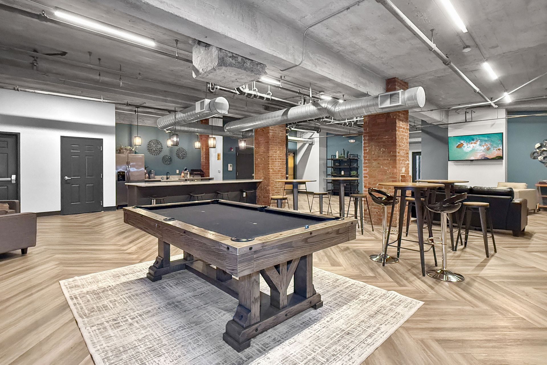 A pool table sits in a modern lounge with industrial-style concrete ceilings, exposed brick, and wooden herringbone floors.