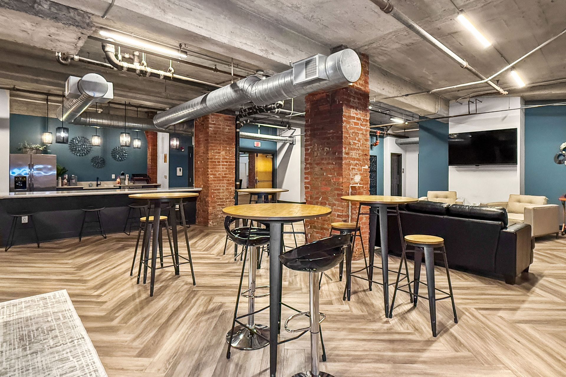 A modern communal space with bar stools, tables, exposed brick pillars, a dark couch, and blue walls on a wood floor.