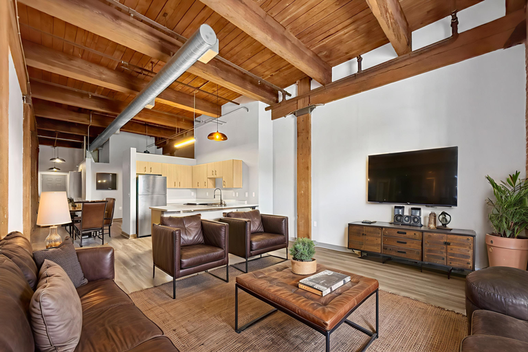 Living room with leather furniture, wooden ceiling beams, a TV, and an open kitchen in the background.
