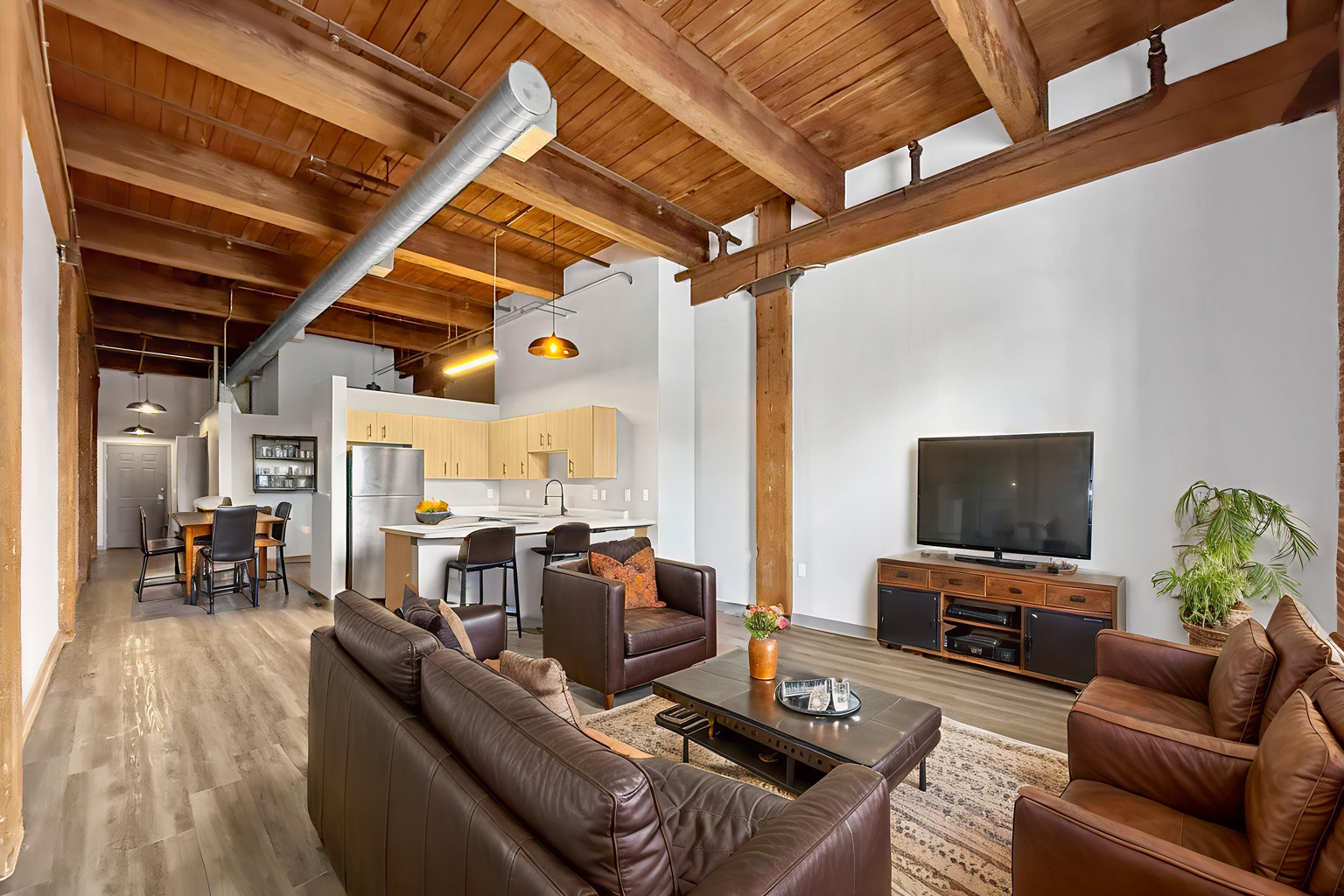 A bright, open-plan loft featuring wooden ceiling beams, a living area with brown leather furniture, and a kitchen.