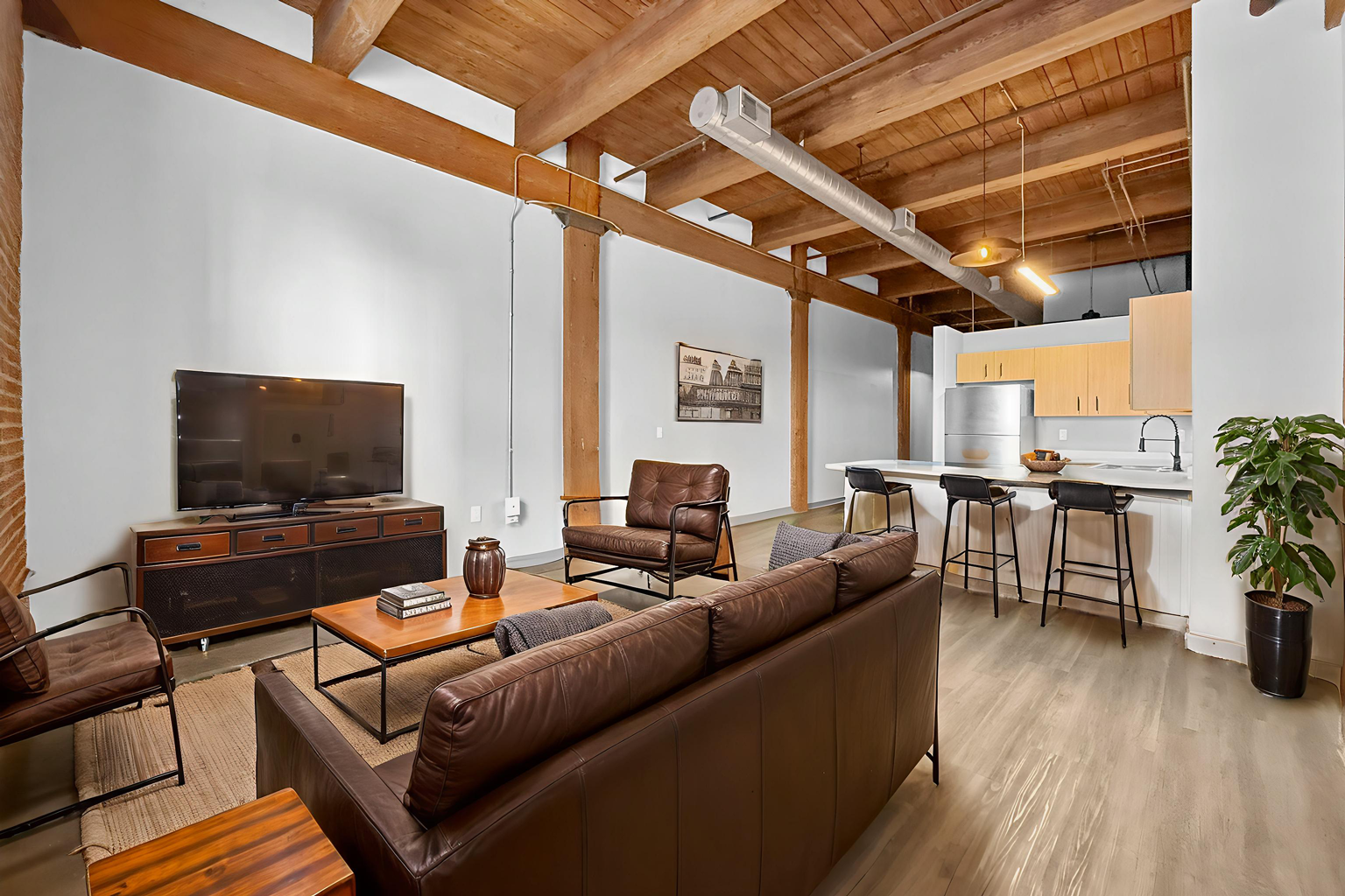 A loft-style living room with a brown leather sofa, wood beams, open kitchen with bar seating, and neutral wall tones.
