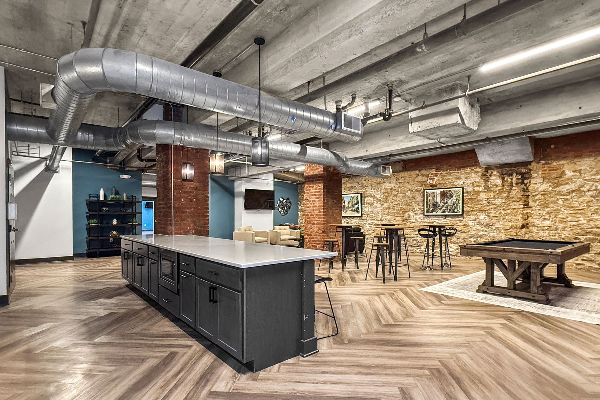 A modern common area featuring an industrial kitchen island, exposed ceiling ducts, brick columns, and a pool table.
