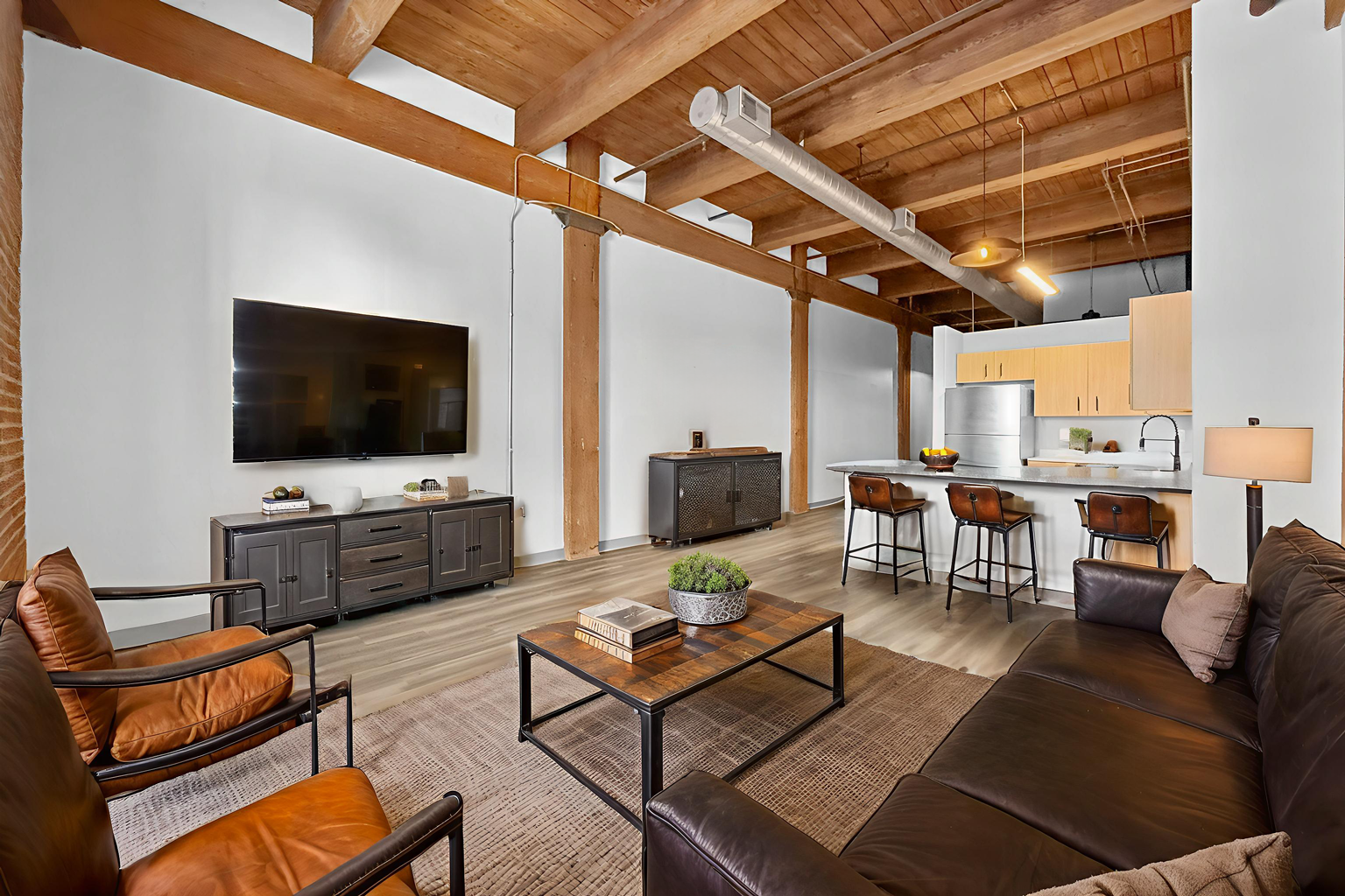 Open-concept apartment with wood-beamed ceiling, leather furniture, industrial decor, and a kitchen with bar seating.