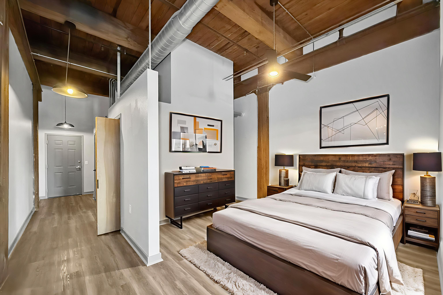 A bedroom with a queen bed, wooden ceiling beams, white walls, and a hallway entry in a modern, industrial-style loft.