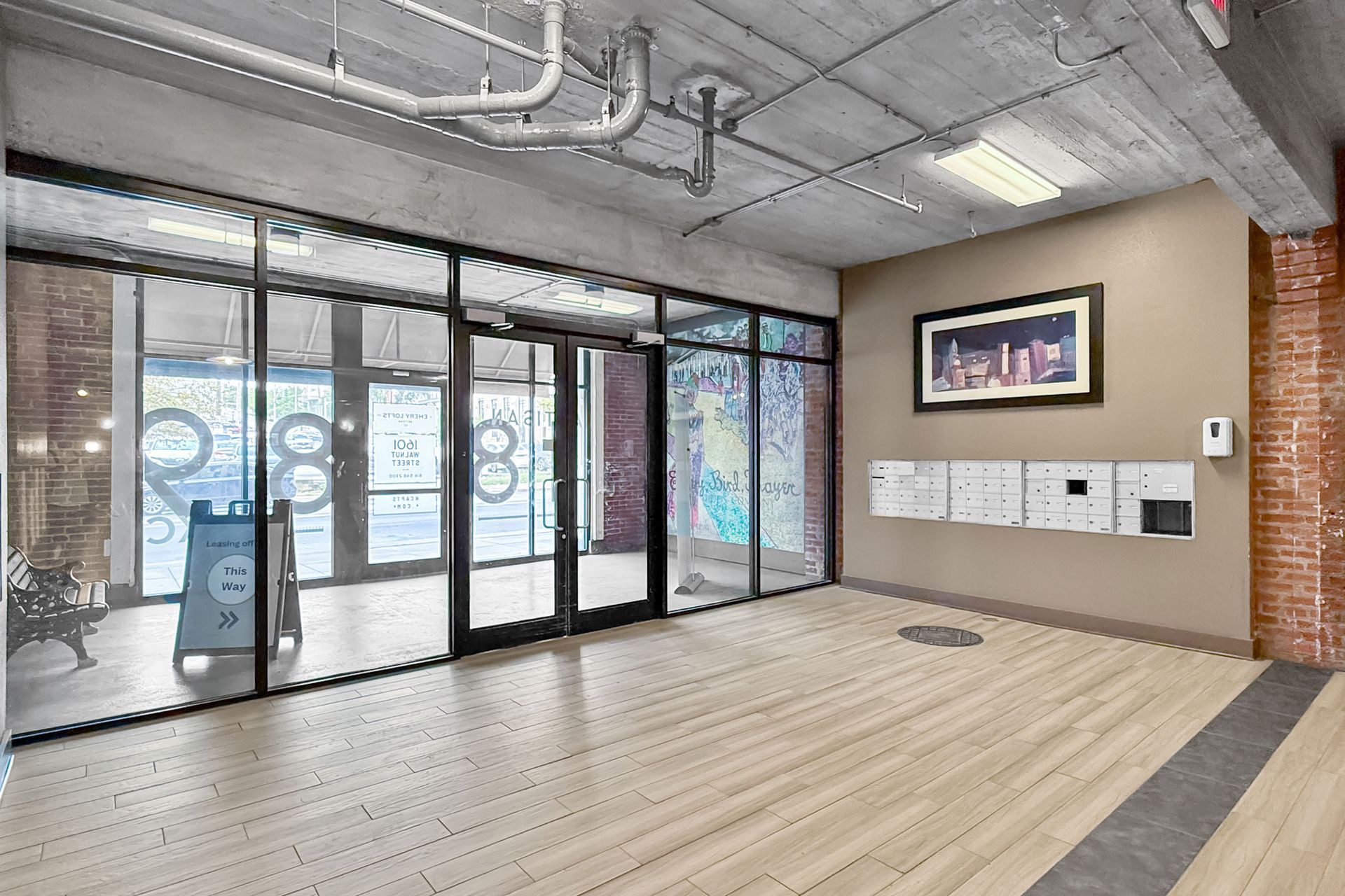 A modern lobby with light wood-look flooring, glass entrance doors, a concrete ceiling, and a wall of mailboxes.