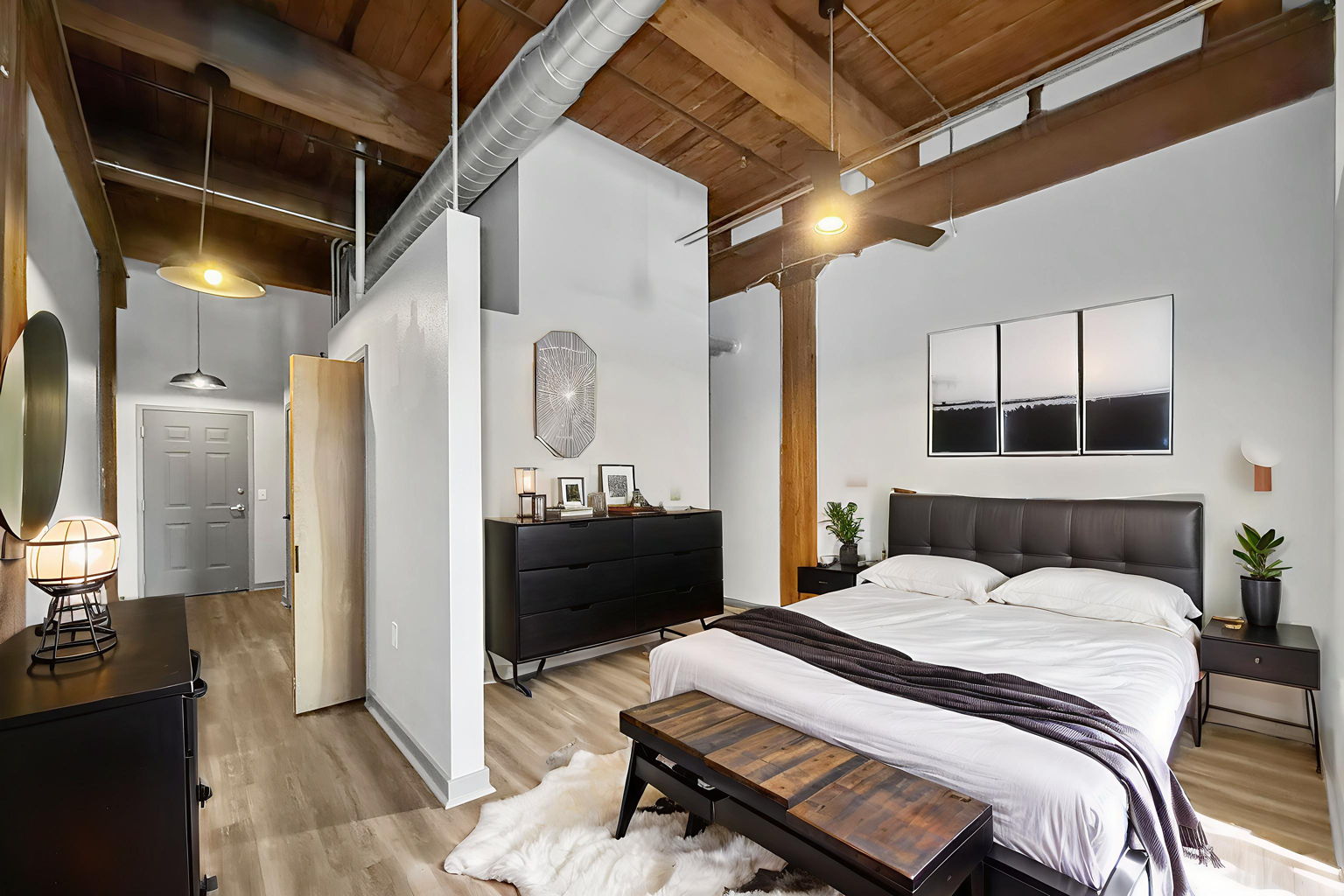 A bright bedroom with wood ceiling beams, a bed with white linens, a dark dresser, and light wood flooring.