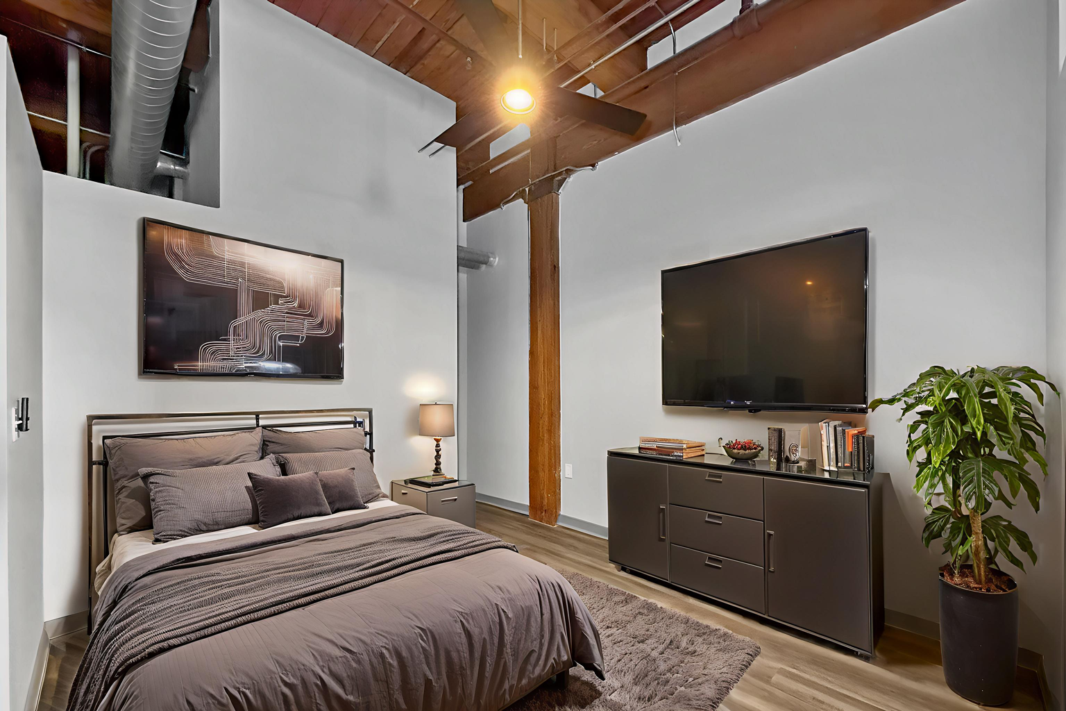 A modern bedroom with high ceilings, exposed wood beams, a gray bed, a flat-screen TV on a dresser, and a potted plant.