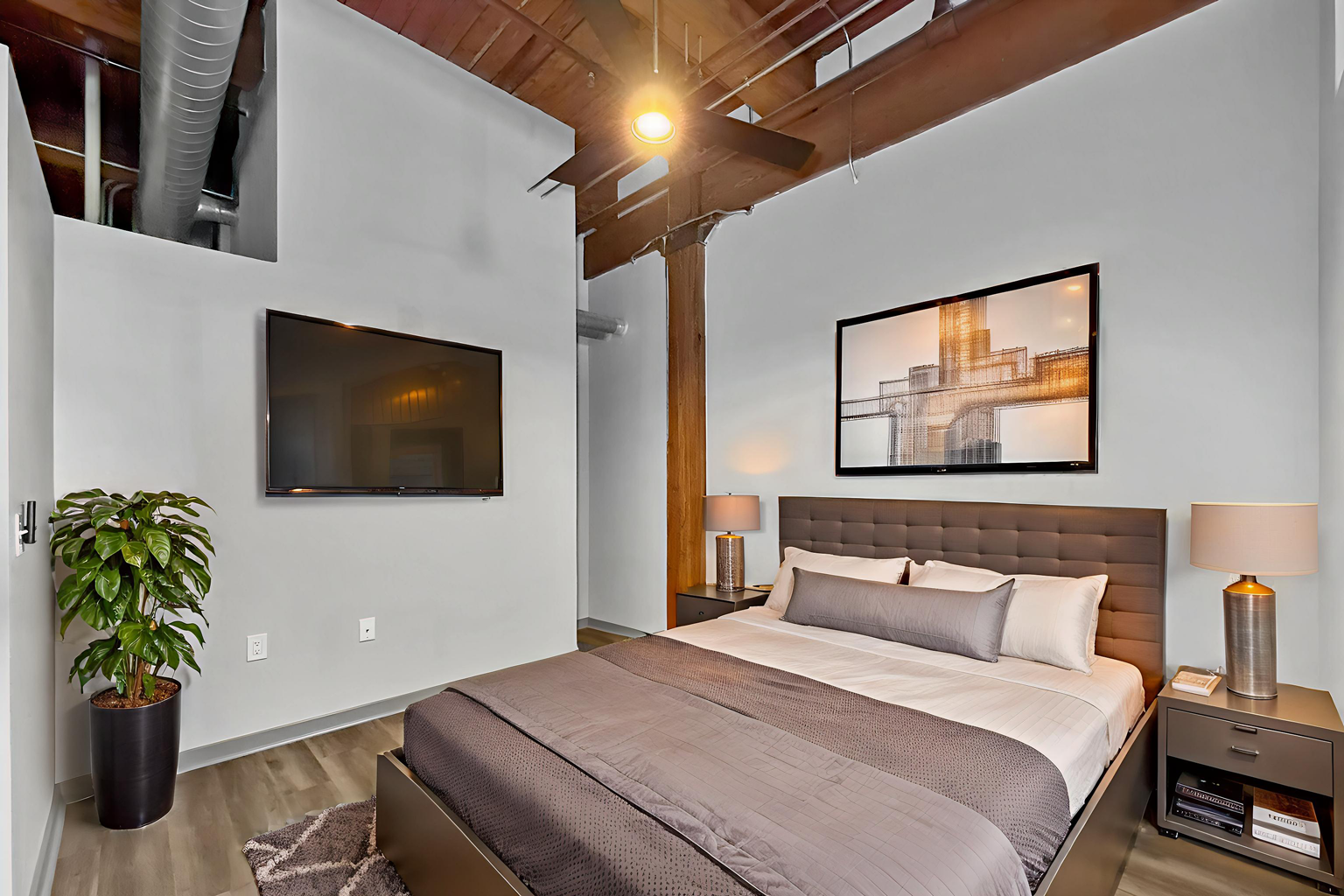 A modern bedroom with high ceilings, a brown upholstered bed, a wall-mounted TV, a potted plant, and a bridge print.