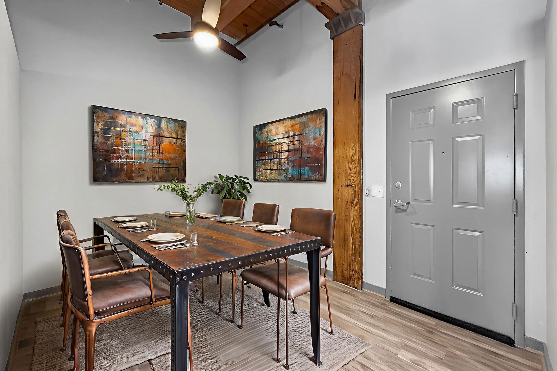 A dining area with a rustic wooden table, six chairs, two abstract paintings, and a grey door in a room with a wood beam.