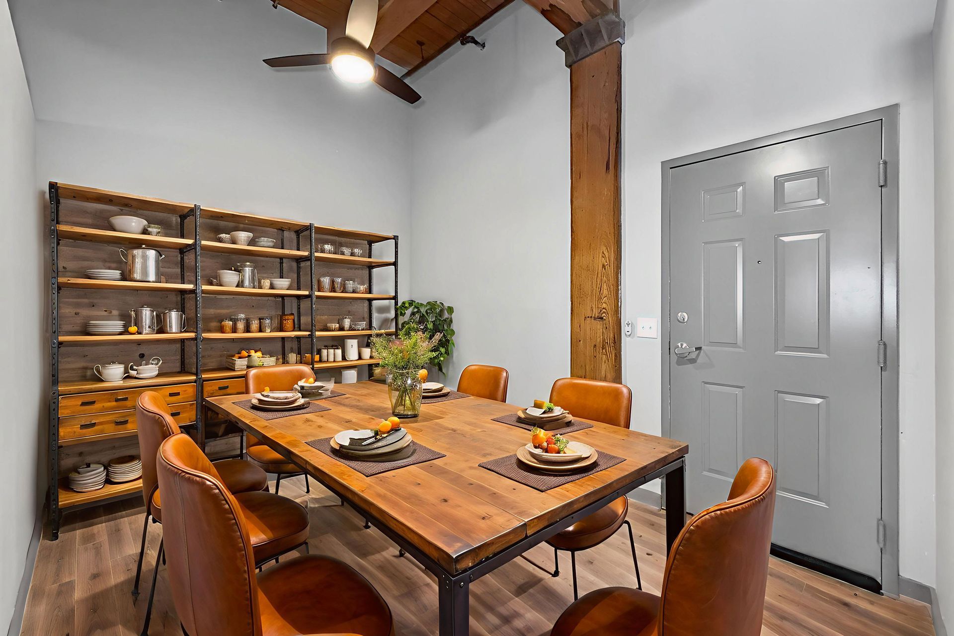 A rustic dining room with a long wooden table, leather chairs, and a large metal shelving unit against a grey wall.