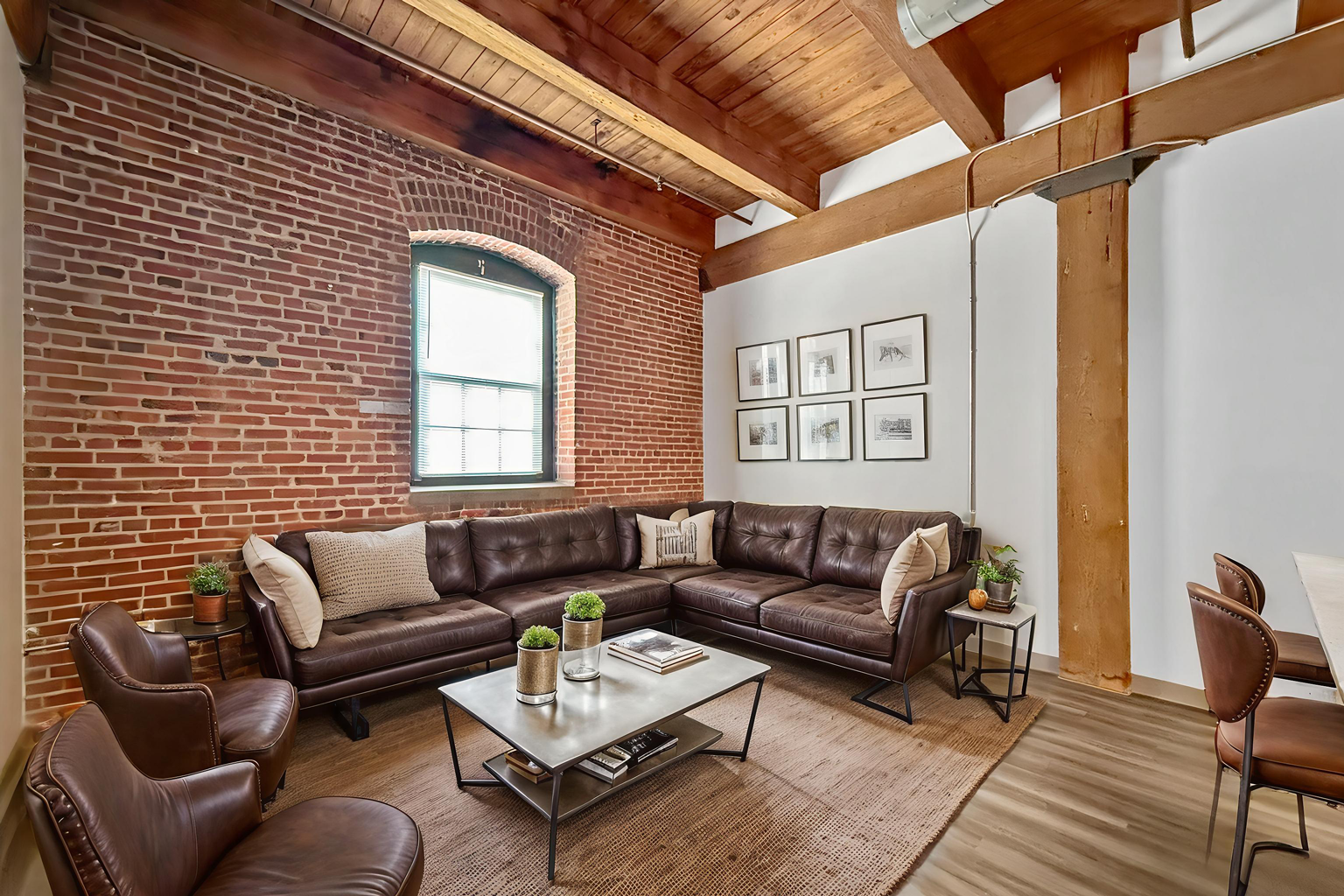 A cozy loft living room featuring a brown leather sectional, two armchairs, a coffee table, and exposed brick walls.