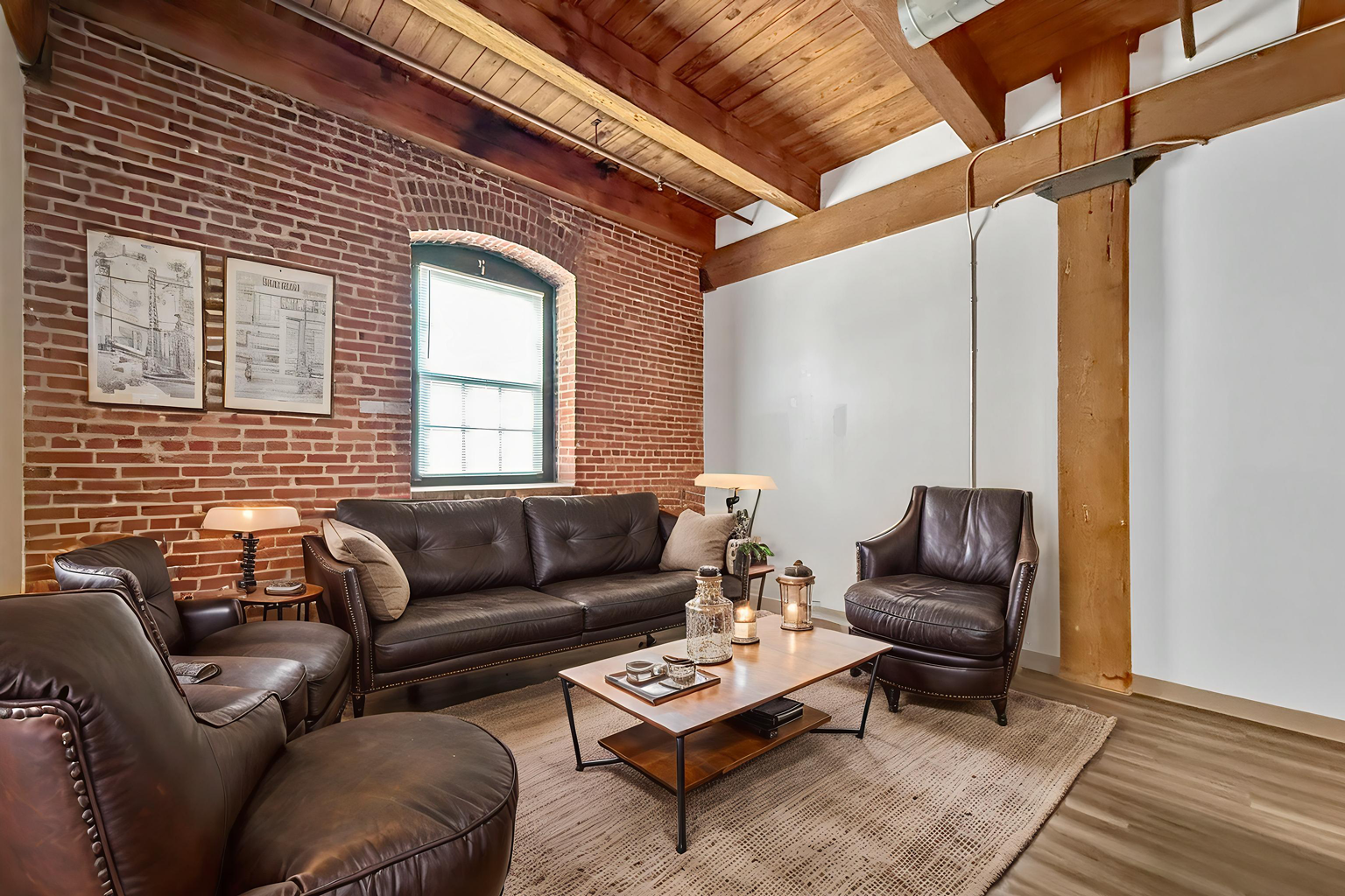 A loft living room with exposed brick walls, timber beams, brown leather furniture, and a central wooden coffee table.