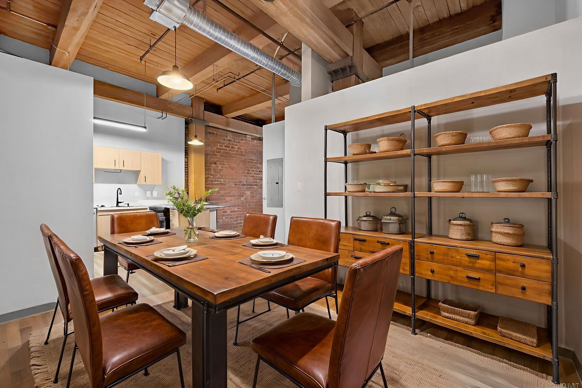 A dining area with a rustic wooden table, brown leather chairs, and a metal shelving unit against a brick wall.