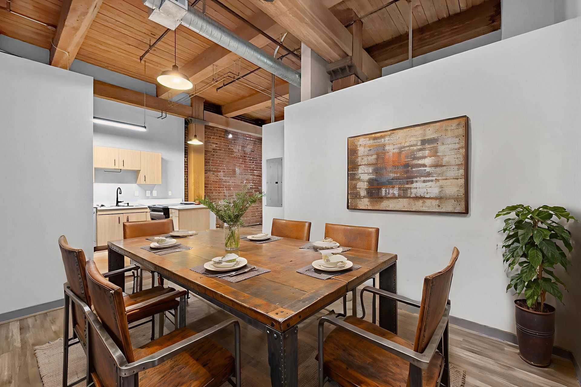 Dining area featuring a rustic wooden table with six leather chairs, an abstract wall art piece, and a kitchen background.