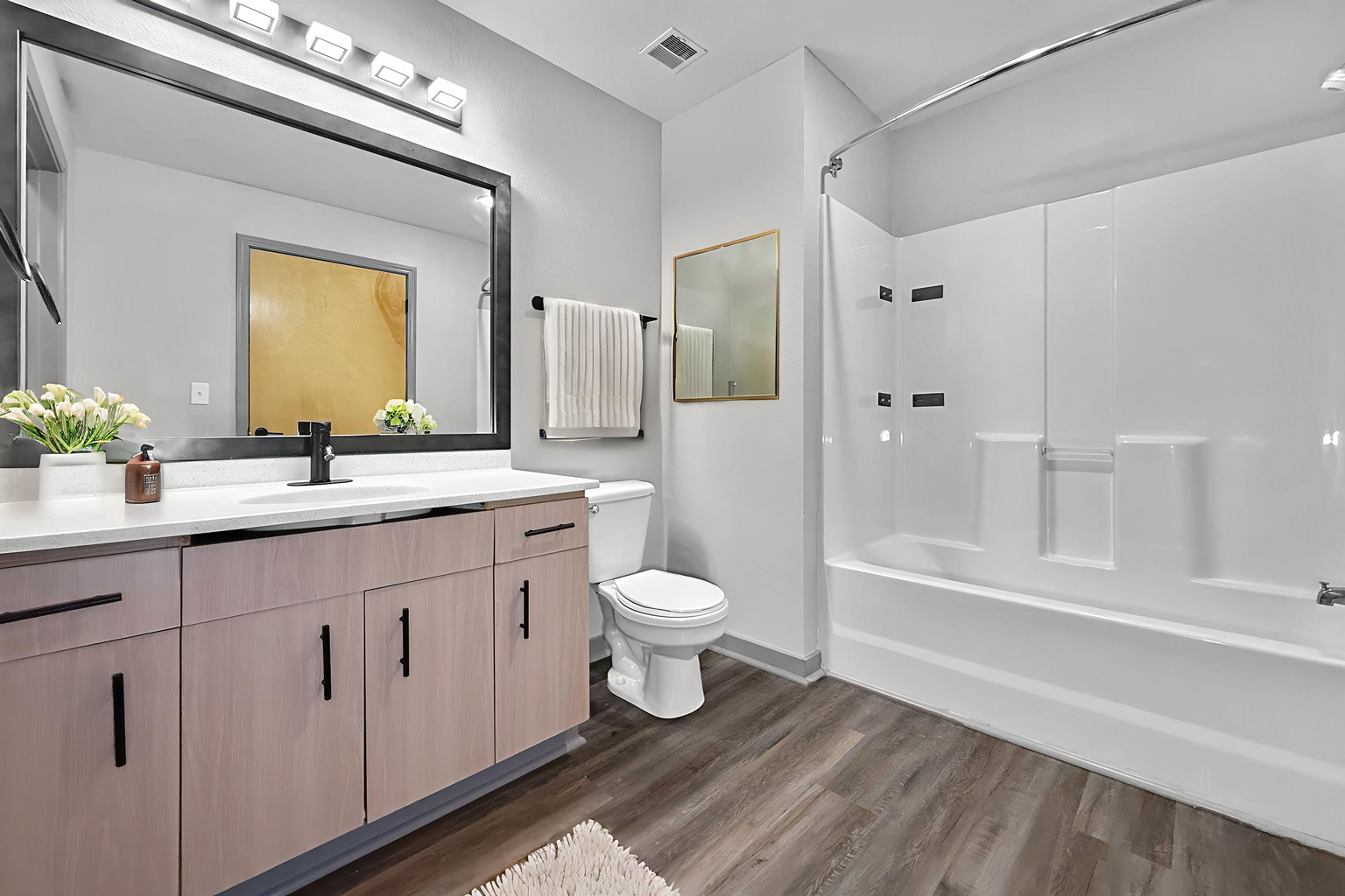Modern bathroom with a wooden vanity, a large mirror, a white toilet, and a bathtub with a white shower enclosure.