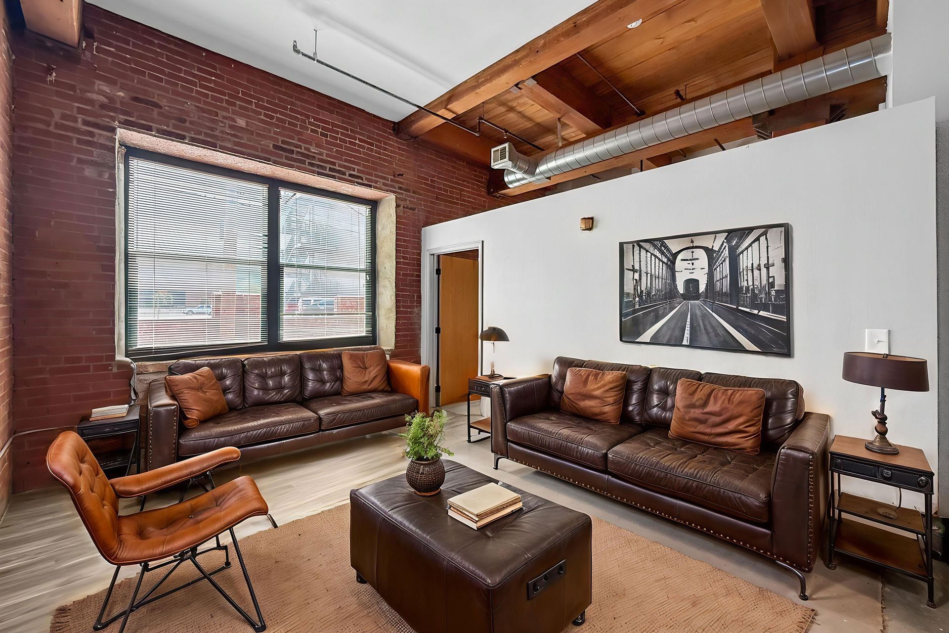 Living room with two leather sofas, a leather ottoman, and a brown armchair against a brick wall under exposed wood beams.