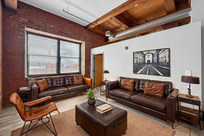 Living room with two leather sofas, a leather ottoman, and a brown armchair against a brick wall under exposed wood beams.