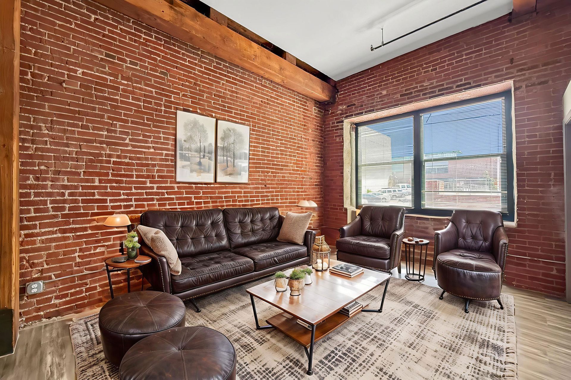 Living room with exposed brick walls, dark leather furniture, a central coffee table, and an area rug under a large window.