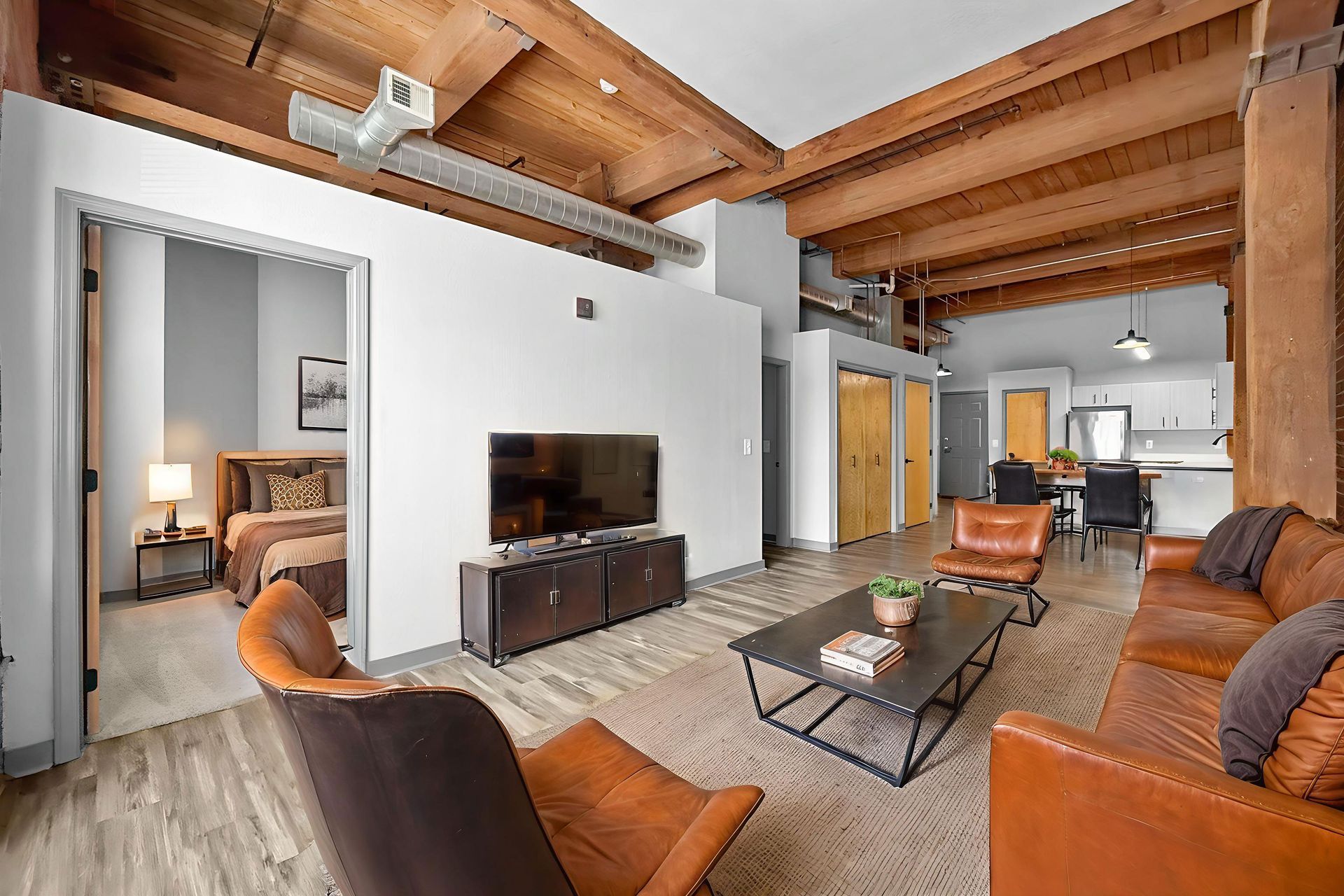 A modern apartment living room with brown leather furniture, exposed wooden beams, and a view into a bedroom.