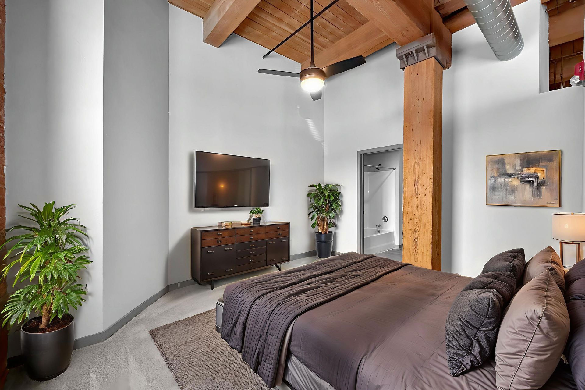 A loft-style bedroom featuring a bed with gray bedding, wooden ceiling beams, a ceiling fan, a TV, and green plants.