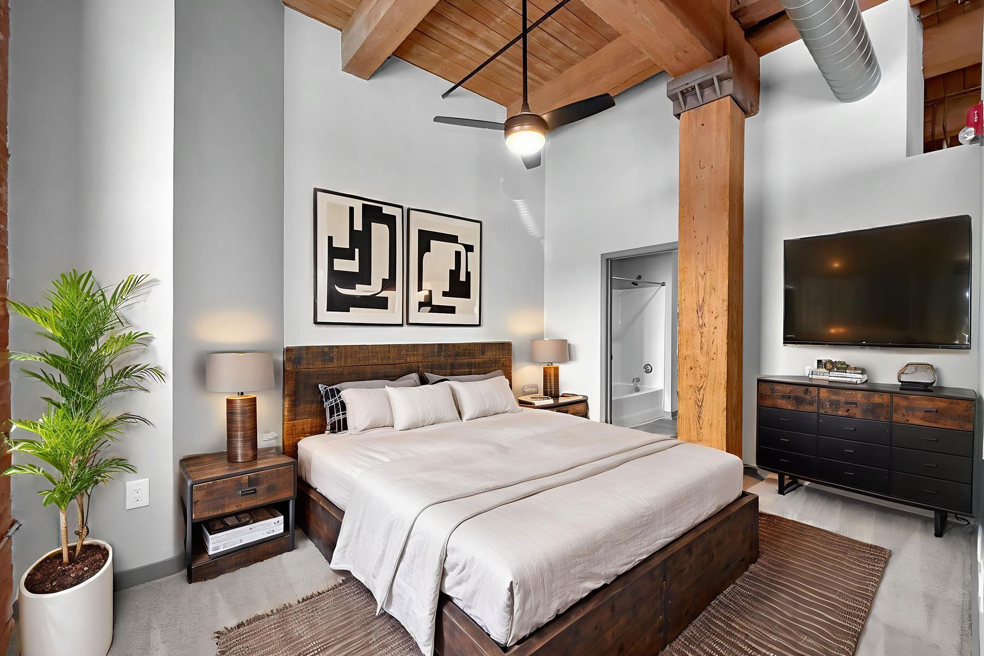 A modern loft bedroom with high ceilings, wooden beams, a platform bed, industrial-style furniture, and a potted plant.