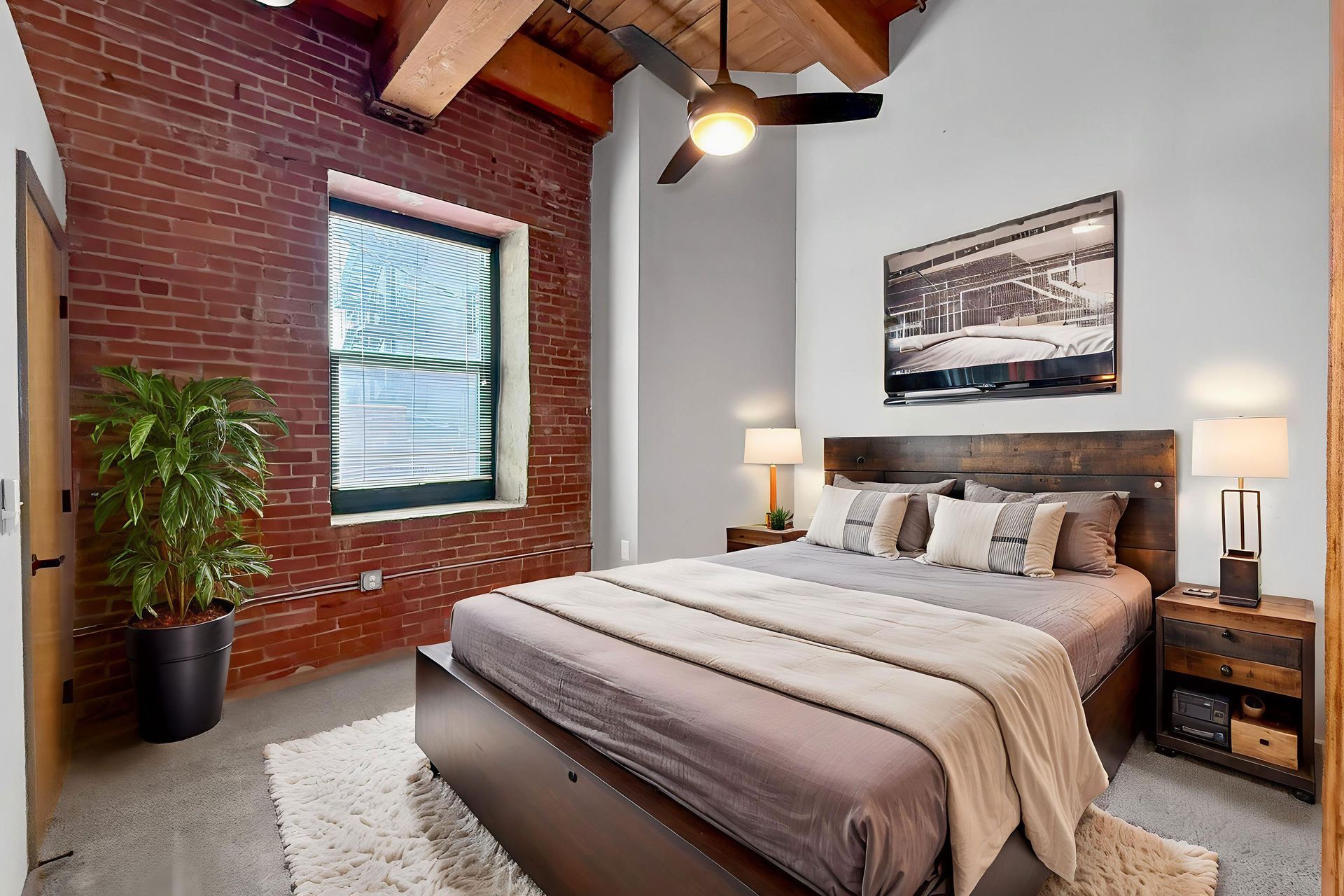 A bedroom with a bed, brick wall, potted plant, wooden ceiling beams, ceiling fan, and bedside tables with lamps.