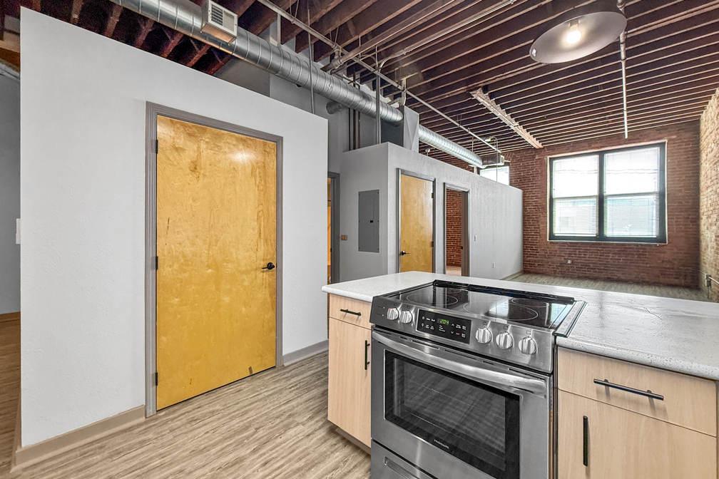 A modern studio apartment interior with a kitchen island featuring a stainless steel stove, facing a brick wall and window.