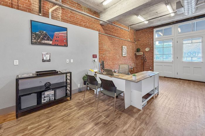 A sunlit office space with exposed brick walls, a modern white desk, two black chairs, and a storage unit on a wood floor.