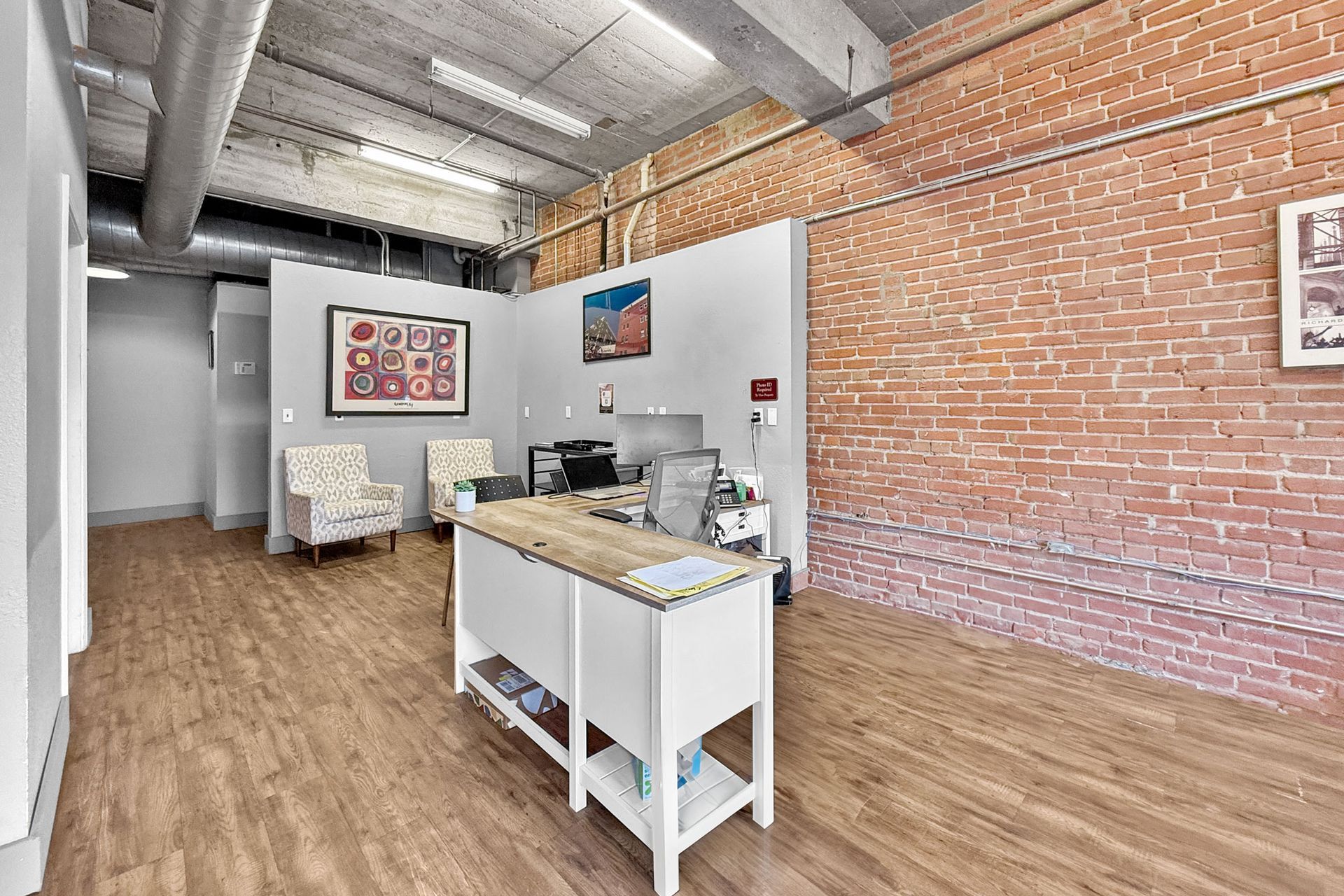 An office reception area with a white desk, two patterned armchairs, wood flooring, and an exposed brick wall.
