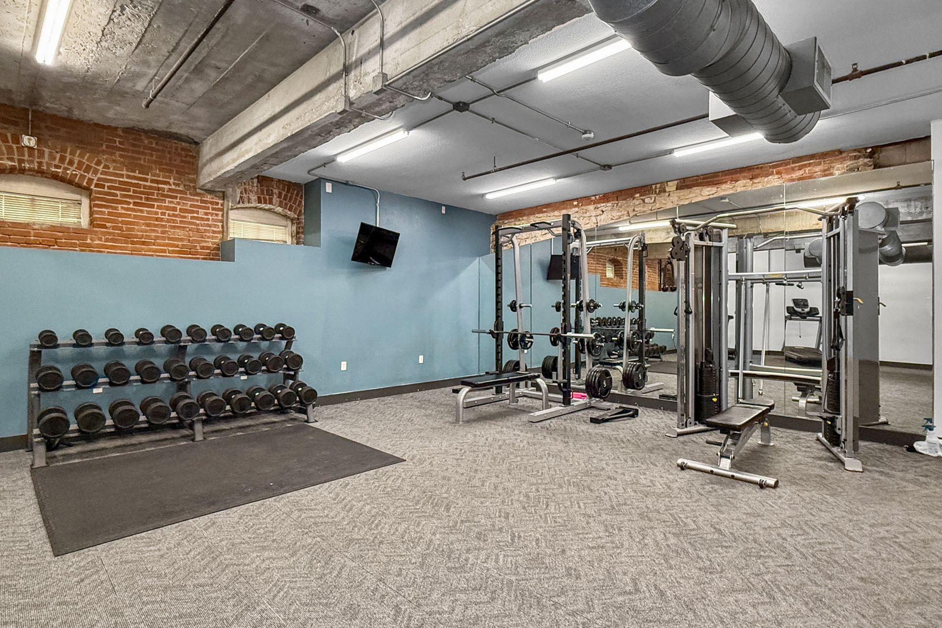 An indoor gym with a dumbbell rack, workout benches, and weight training equipment against a brick and blue wall.